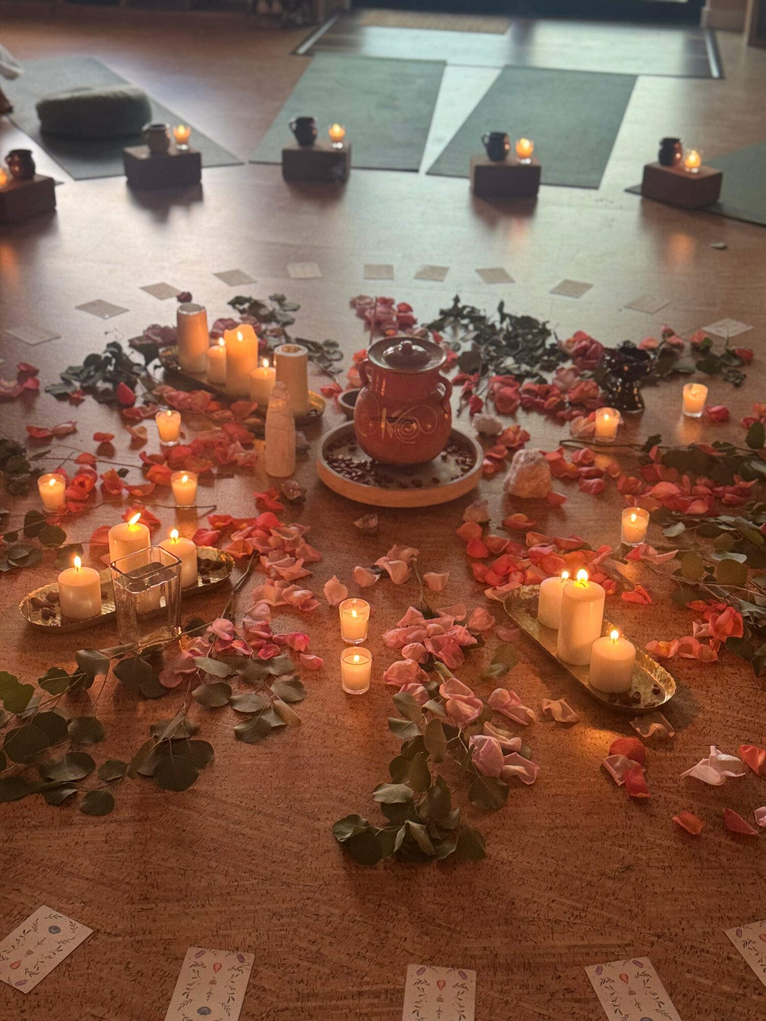 Decorative arrangement with candles, rose petals, and a central object on a wooden surface, with a tiled floor in the background.