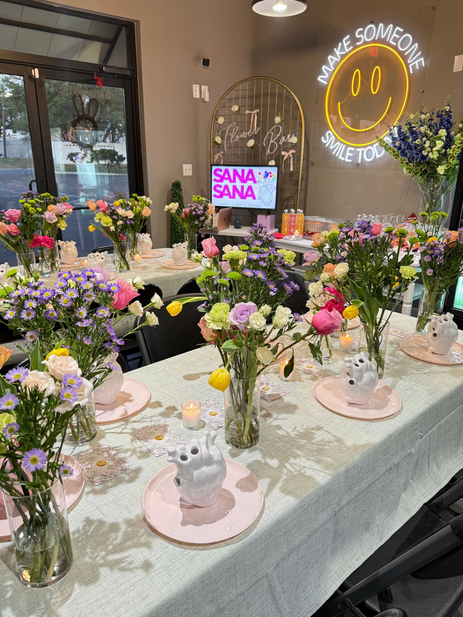 Table with colorful flower arrangements, a cake, and a sign that says 'SANA SANA' in a decorated indoor space.