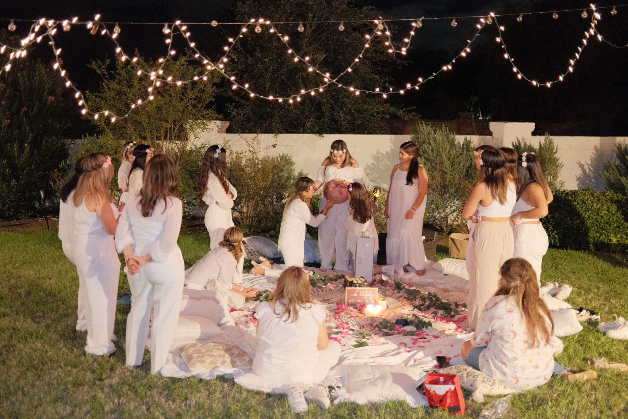 Group of people dressed in white gathered outdoors under string lights, sitting and standing on a blanket, participating in a ceremony.