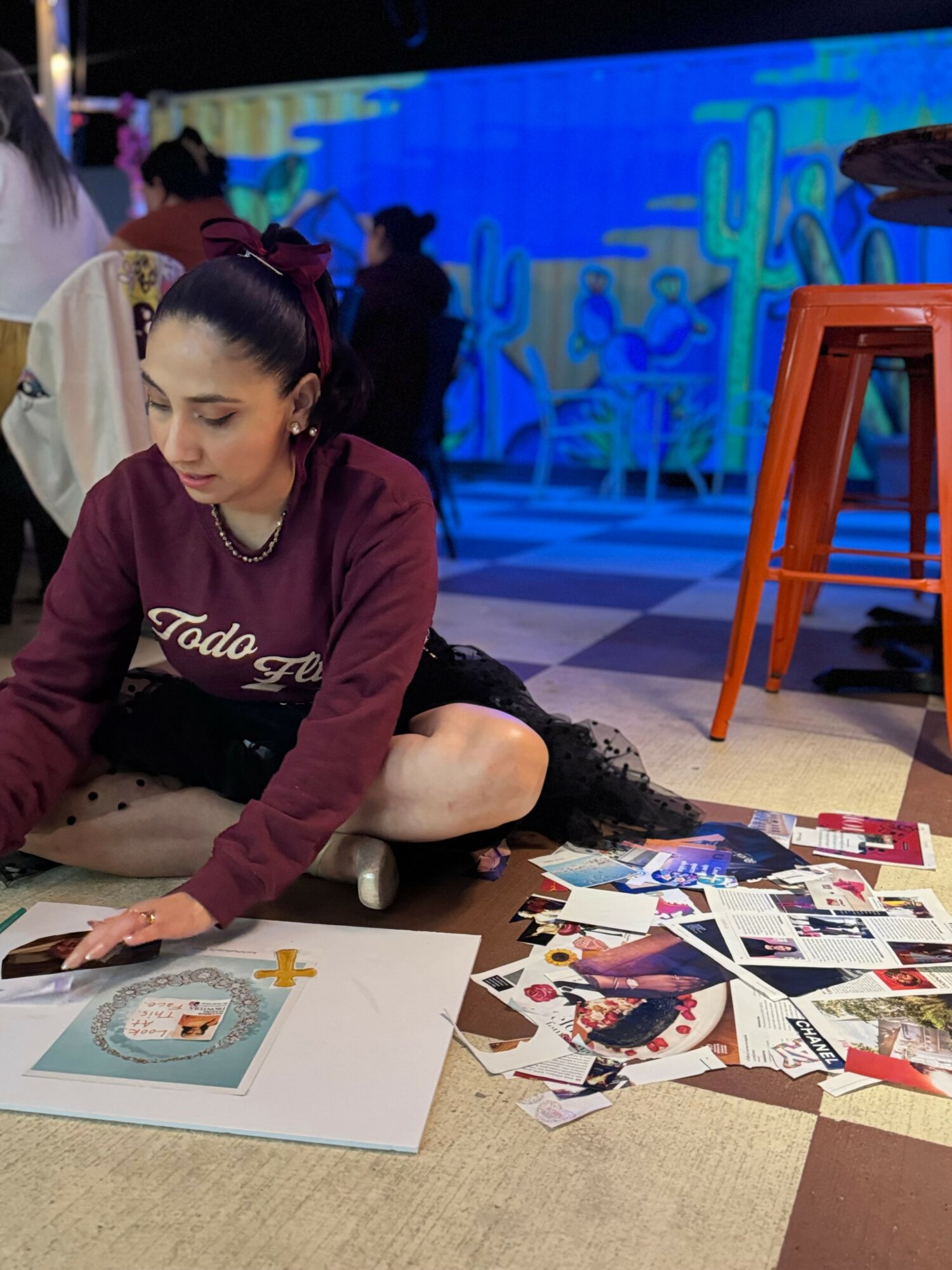 Woman sitting on floor arranging photographs and artwork, with colorful background and orange stool nearby.
