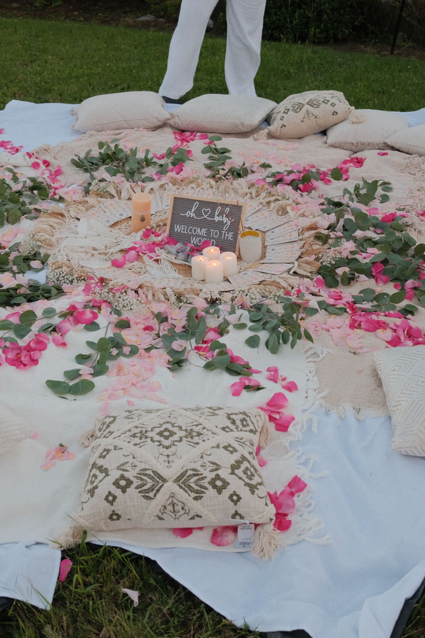 Outdoor wedding setup with a decorated floral arrangement, candles, pillows, and a small chalkboard sign on a white cloth.