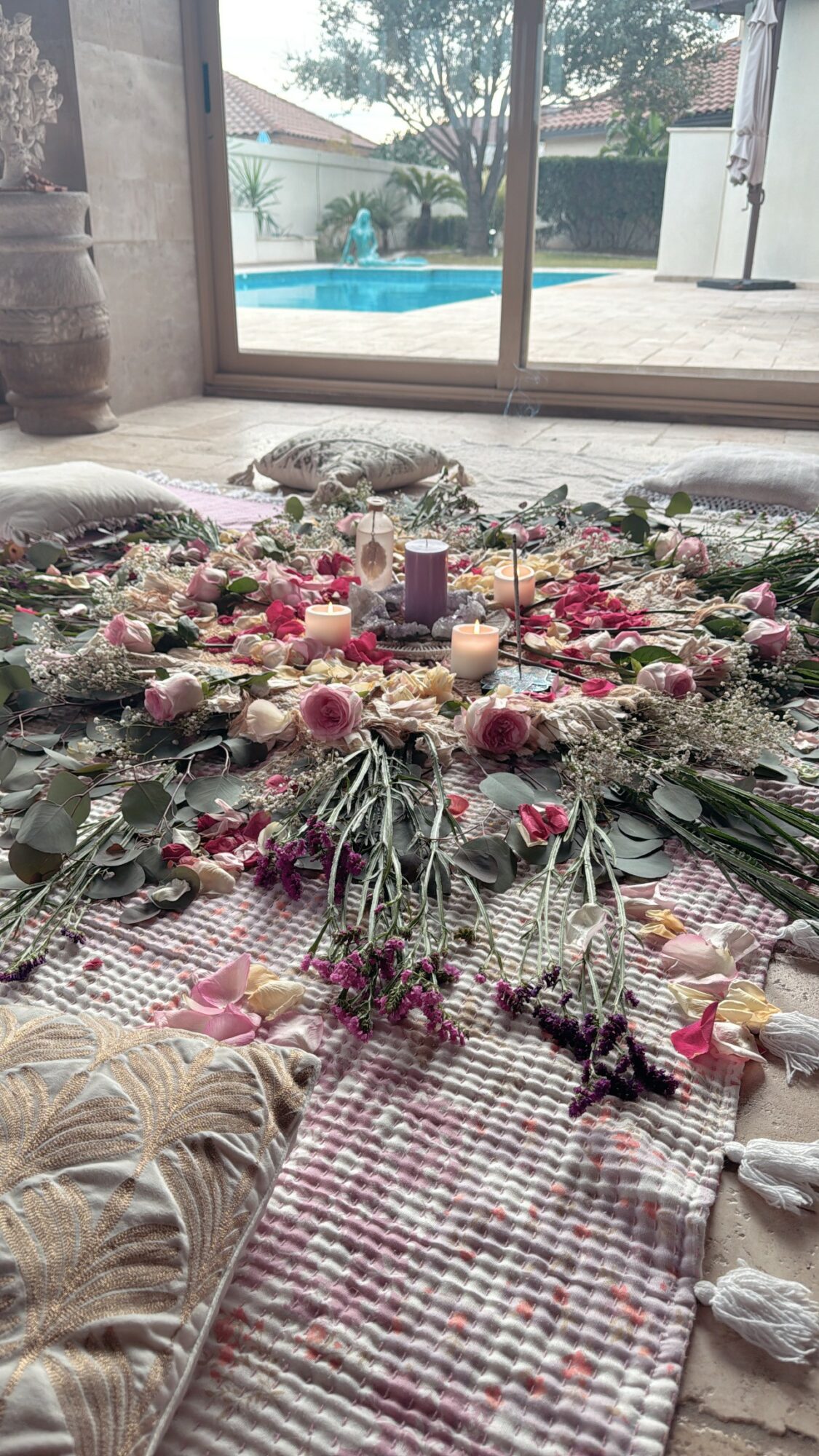 Decorative arrangement with flowers, candles, and greenery on a patterned cloth near a window with a pool outside.