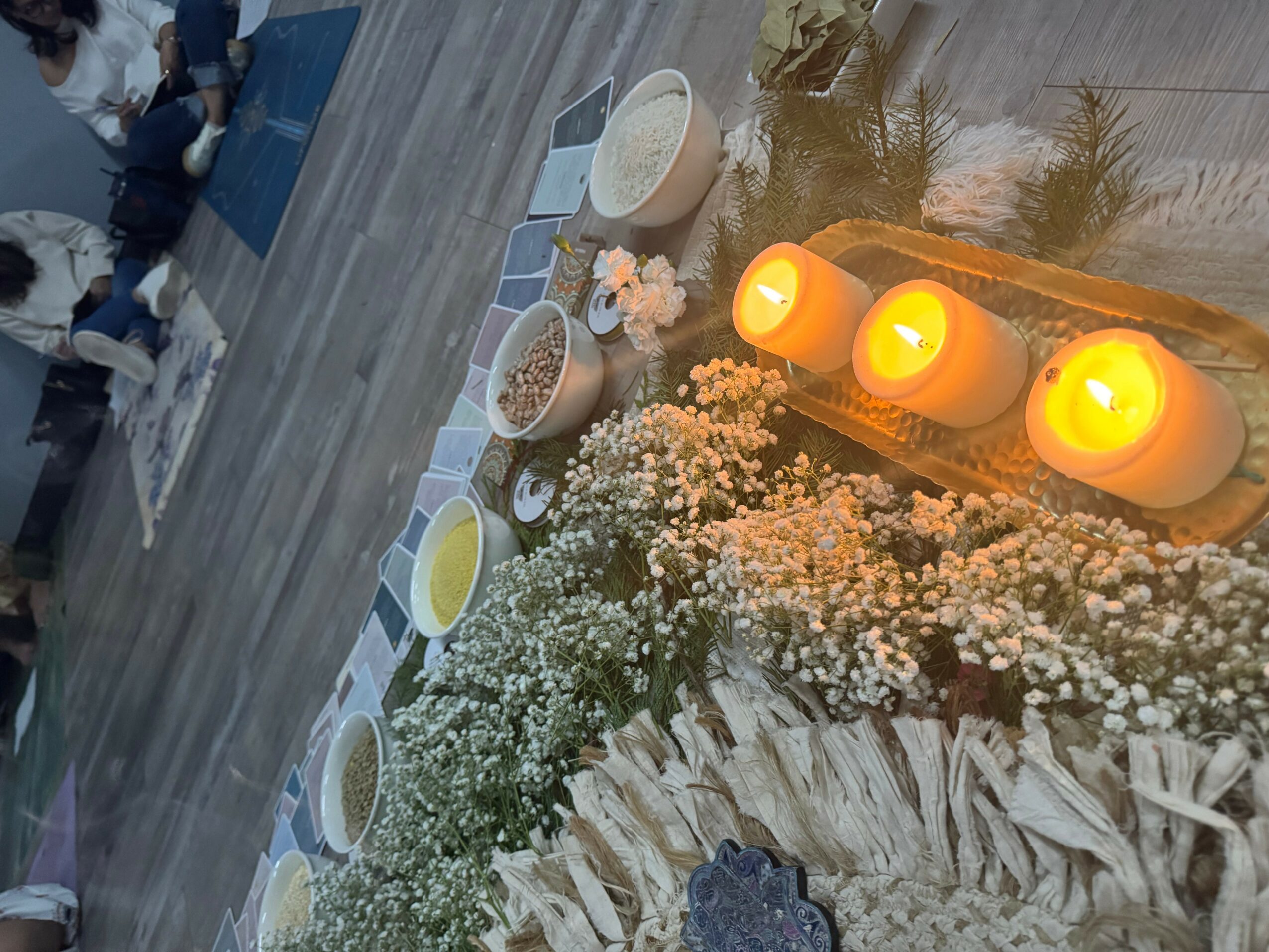 Decorative candles, flowers, and bowls with grains on a table, with a reflection of plants and a window, in a cozy setting.