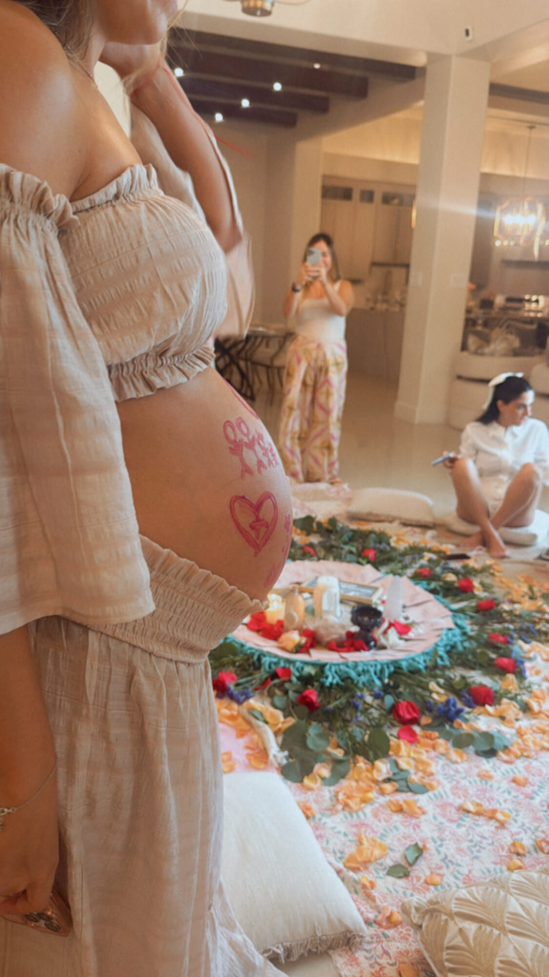 Person with a pregnant belly decorated with pink hearts, standing near a festive display on the floor, in a well-lit room.