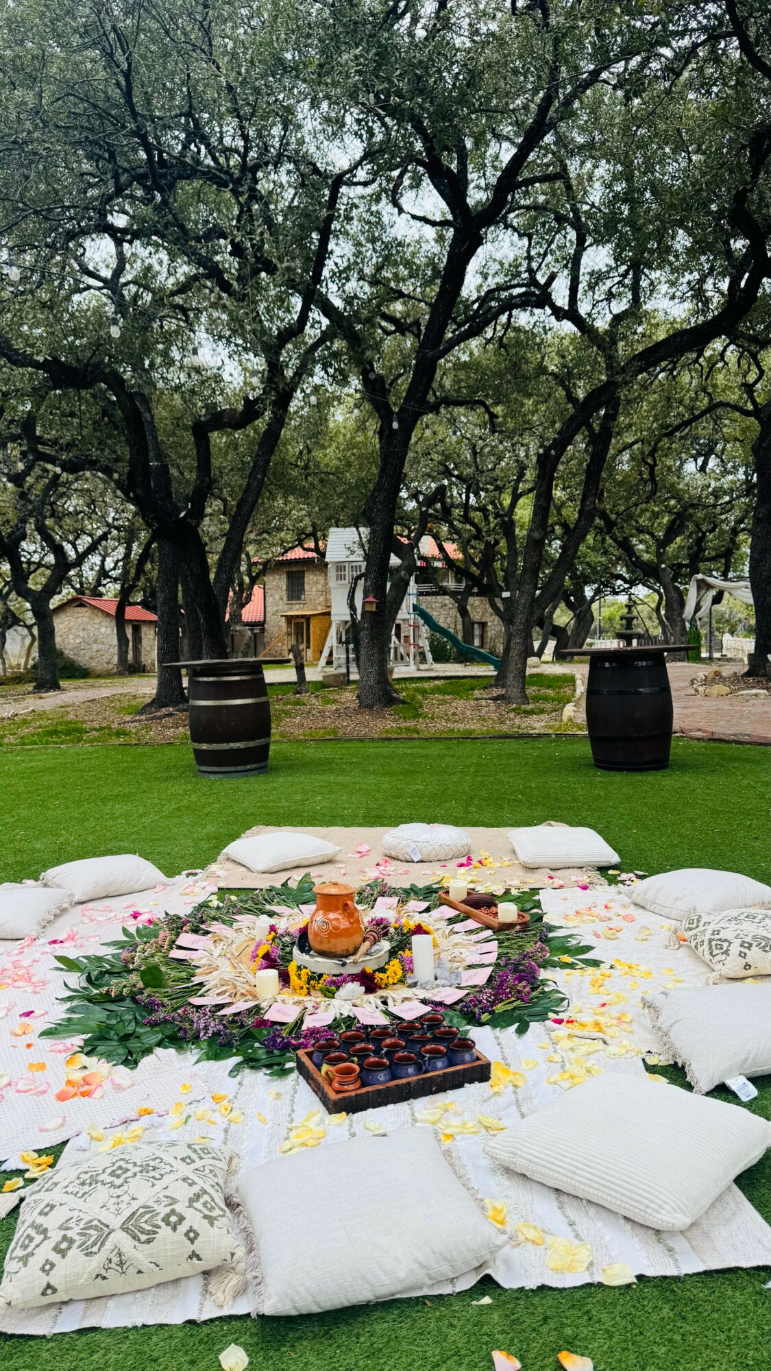 Outdoor scene with a colorful flower arrangement on white cushions, trees, barrels, and a building in the background.