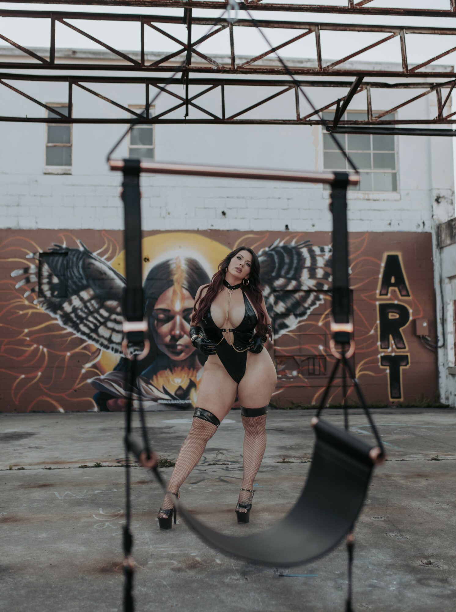 Woman in black lingerie and high heels standing in front of a butterfly mural, with a swing in the foreground.