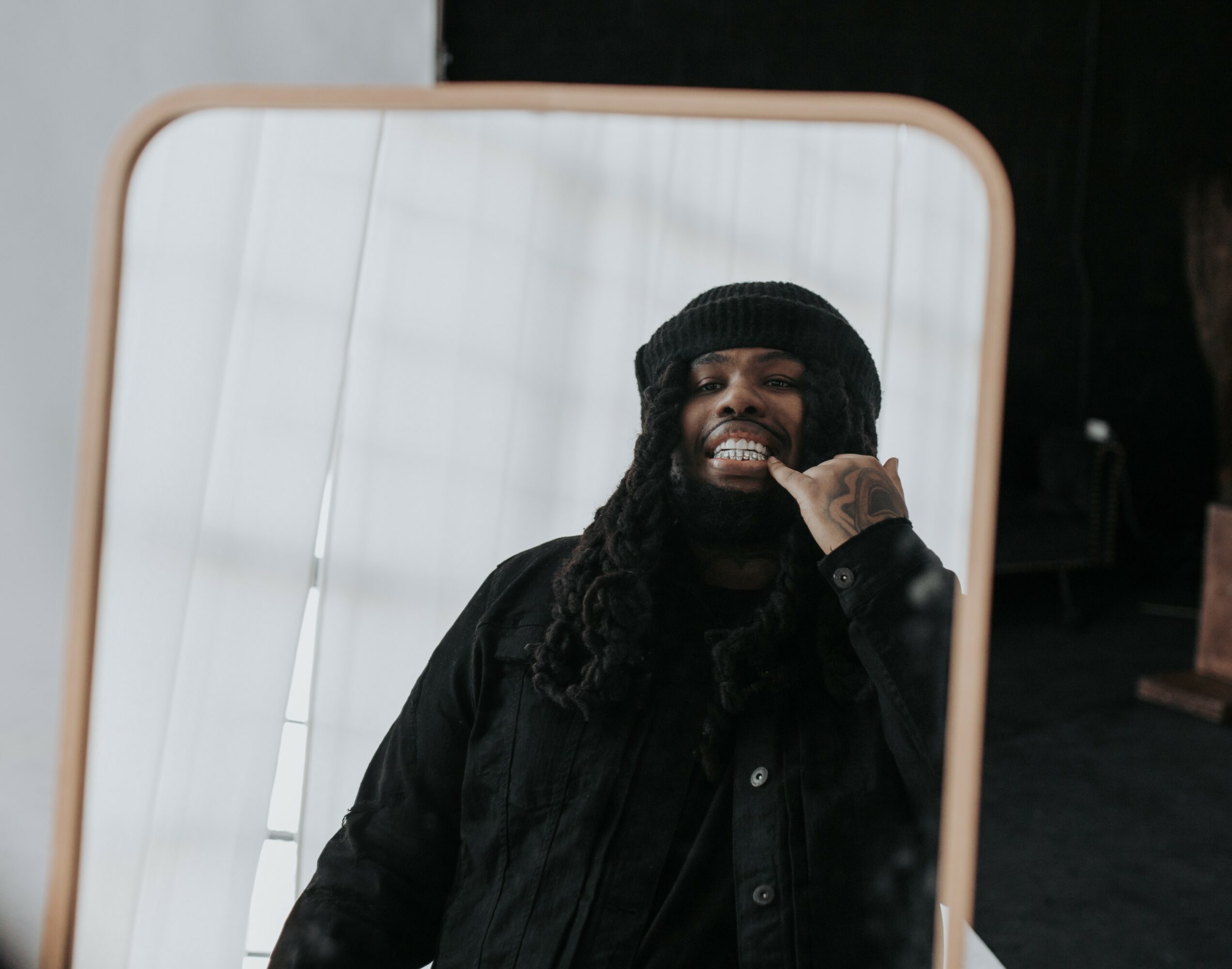 Person smiling and making a hand gesture in front of a mirror, wearing a black beanie and jacket.