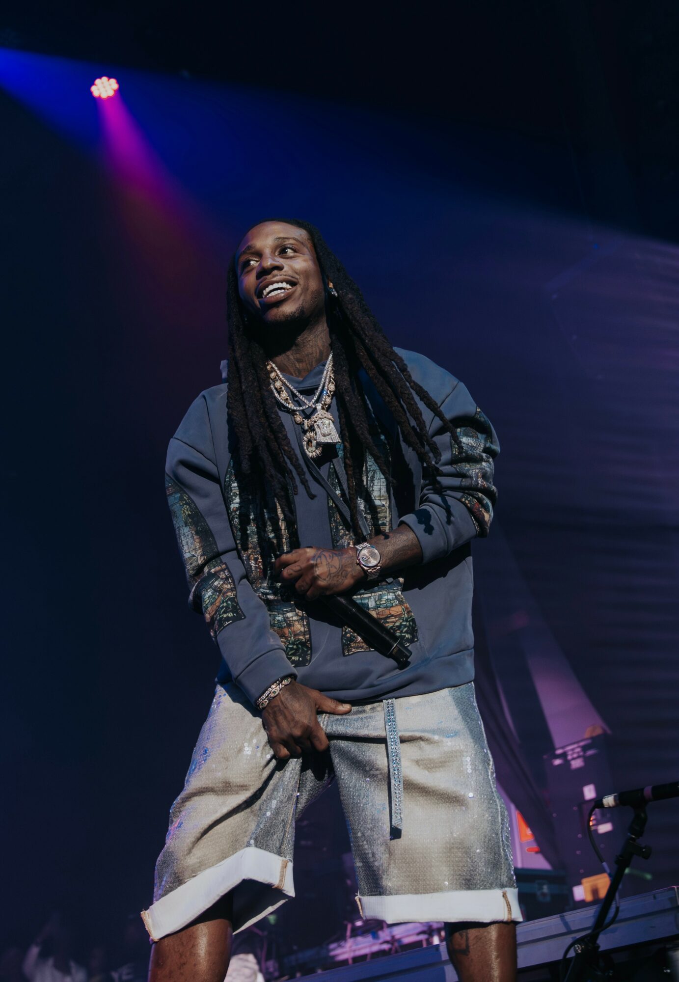 Person with long dreadlocks smiling on stage, wearing a dark jacket and light-colored shorts, with stage lighting in background.