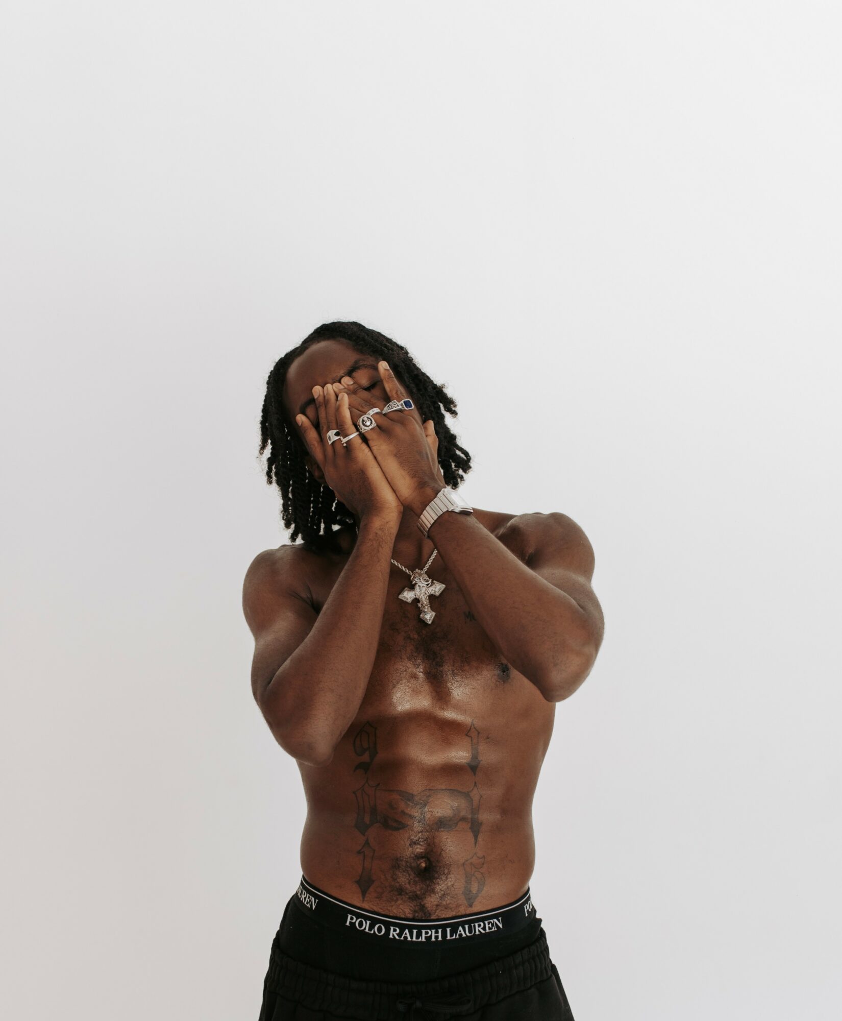 A shirtless person with tattoos covering their torso, covering their face with hands, against a plain white background.