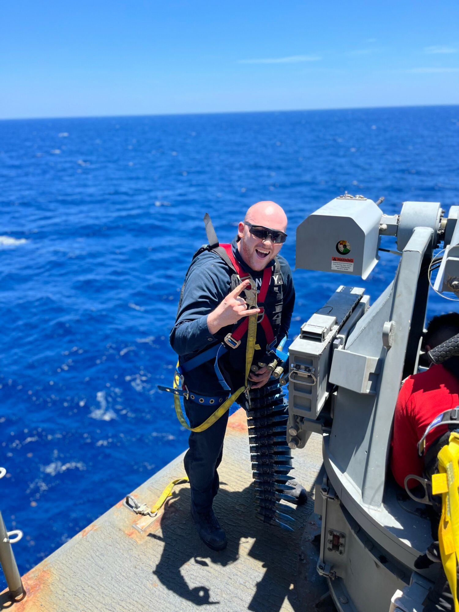 Person in sunglasses and jacket on boat, smiling, making a peace sign, with ocean background.