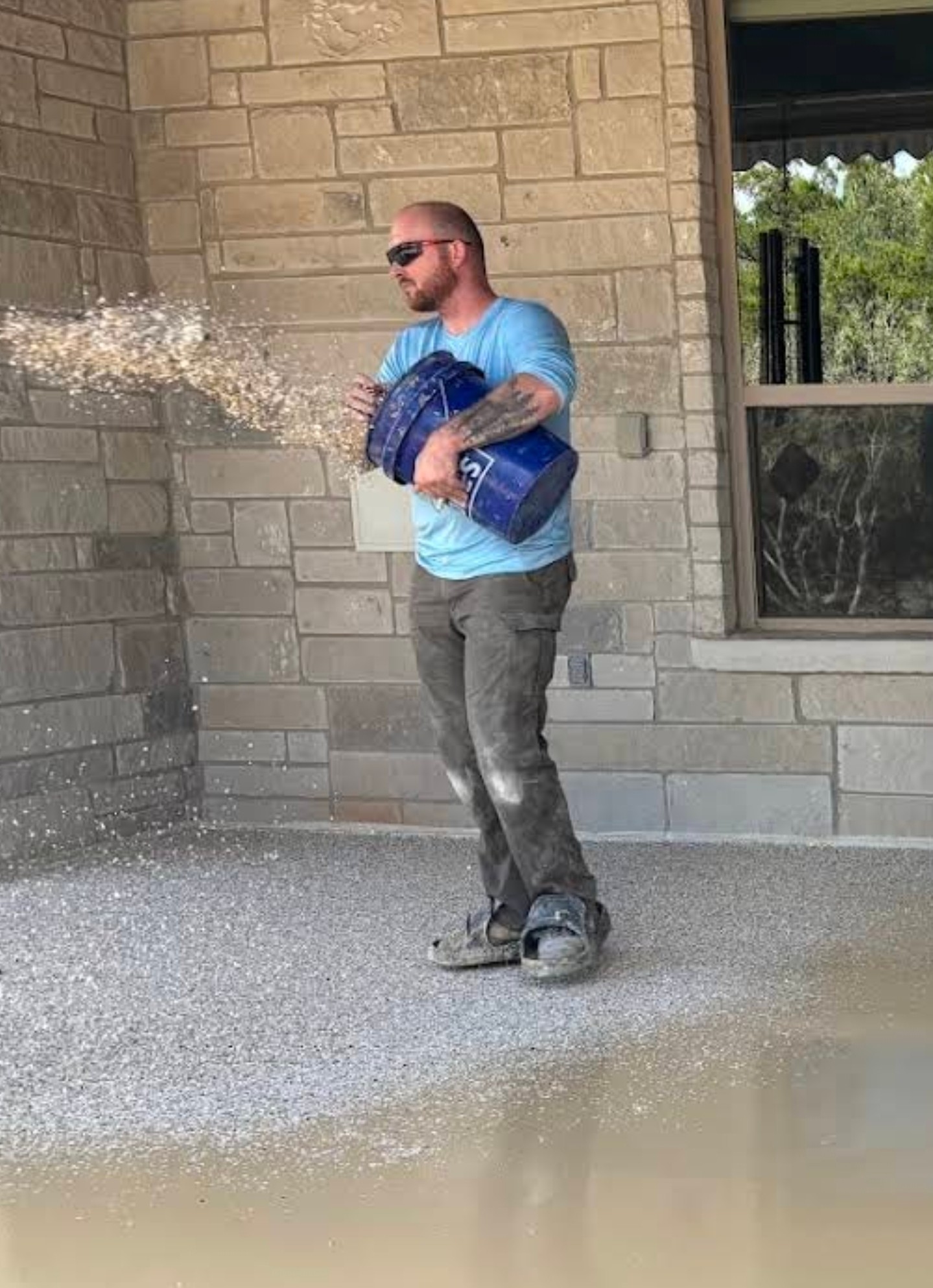 Person wearing sunglasses and gloves, holding a container, spraying a substance outside a building with brick walls and a window.