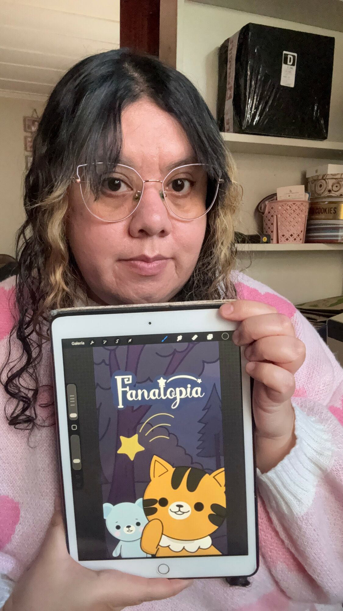 Person with glasses holding a tablet displaying a colorful cartoon with a tiger, a star, and a cat, in a room with shelves.