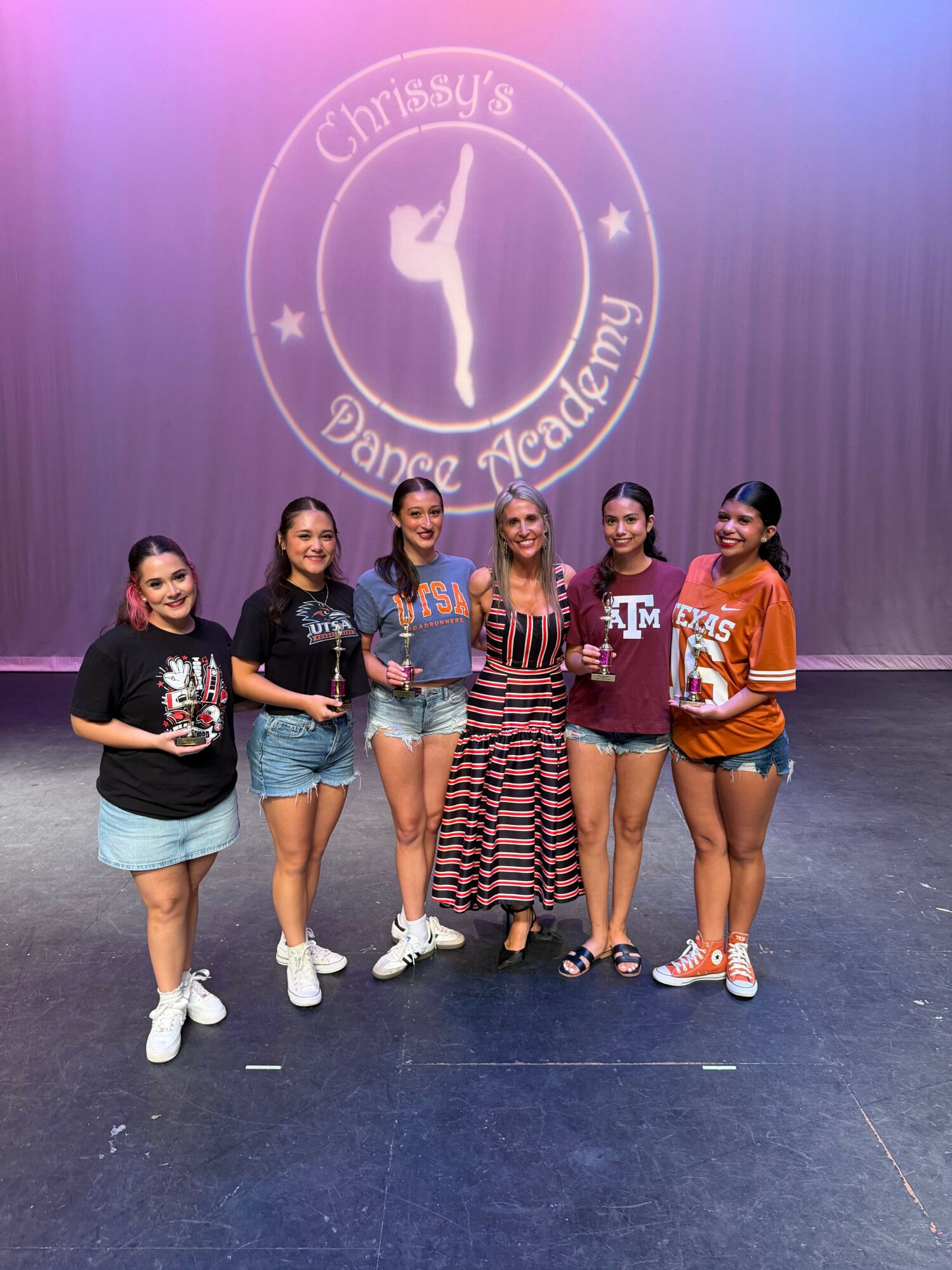 Five young women stand together in front of a purple backdrop with a dance studio logo, smiling and posing casually.