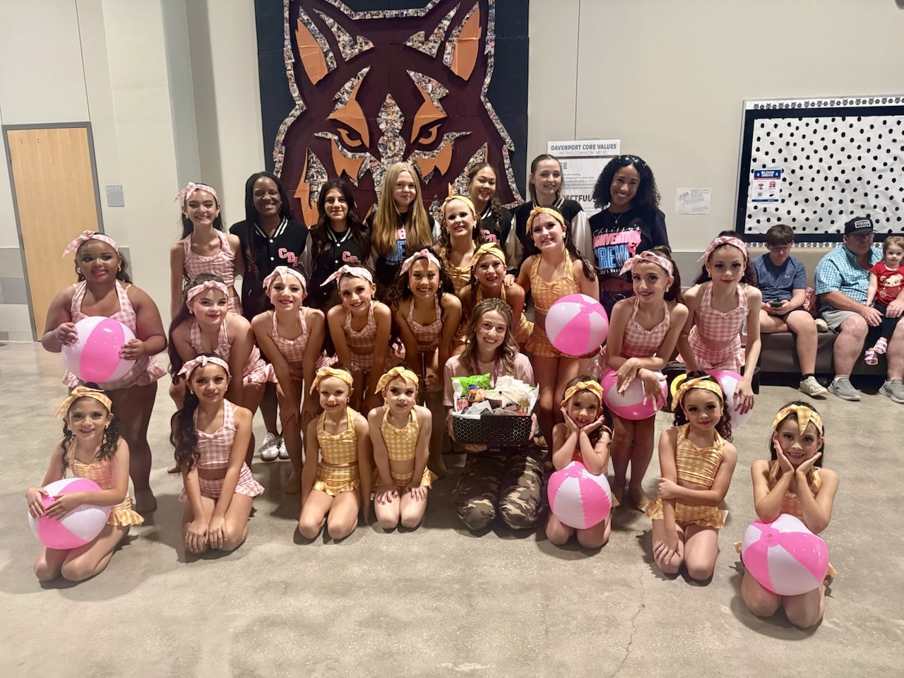 Group of young girls in dance costumes posing with a cake and pink balloons, indoors with a wolf tapestry in background.