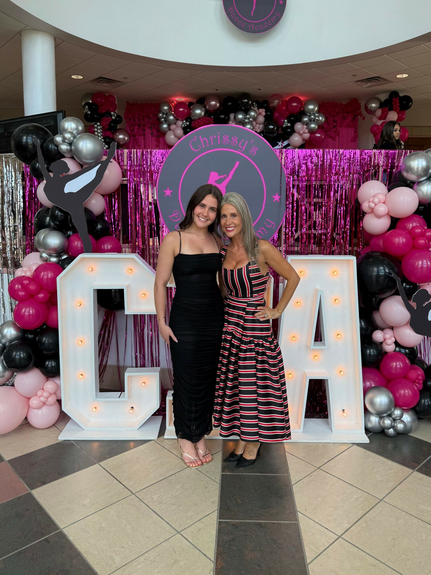 Two women stand in front of large illuminated letters C and A, with pink, black, and silver balloons and a pink backdrop behind them.