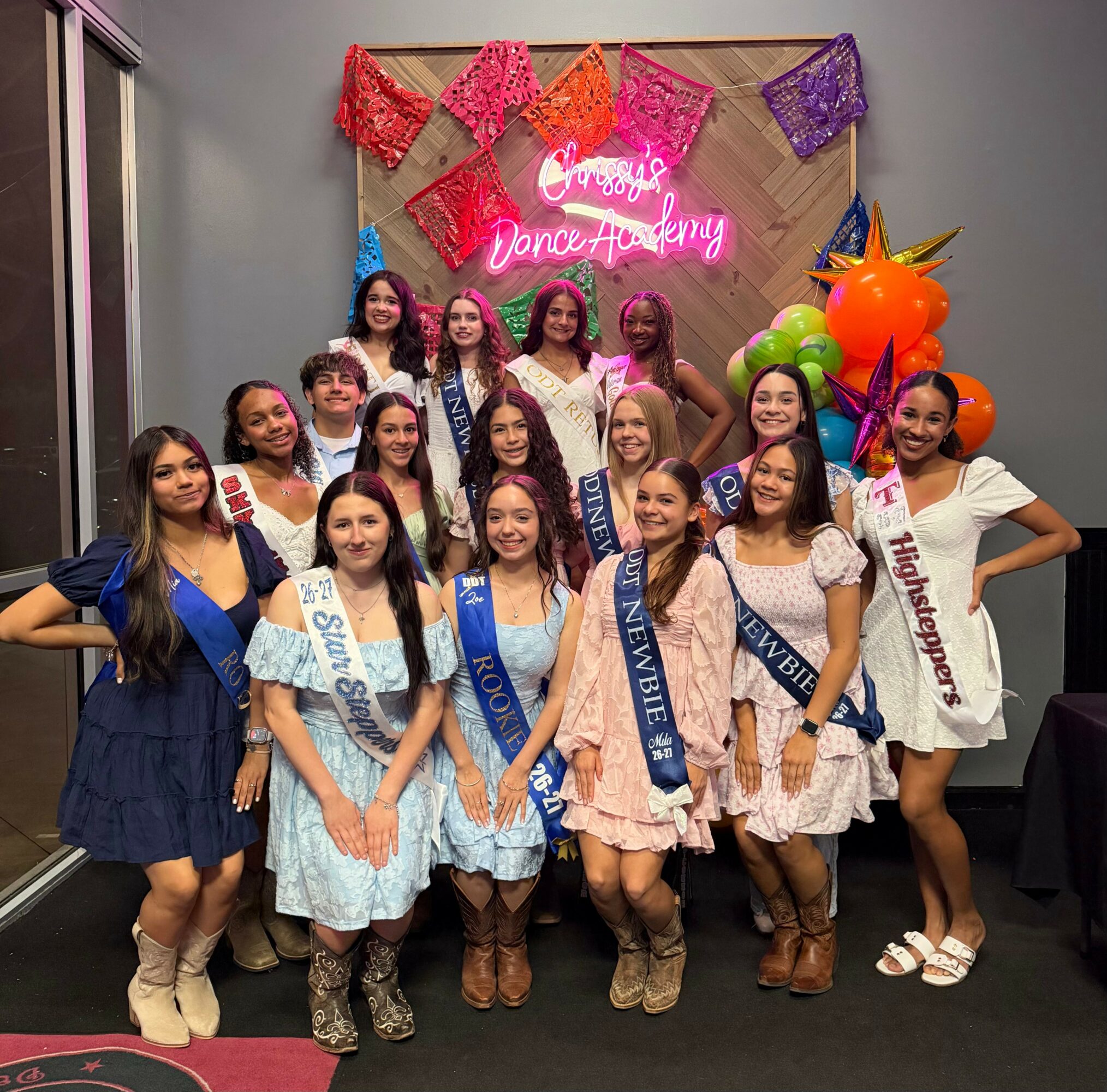 Group of young women in dresses and sashes posing indoors with colorful decorations and a neon sign background.