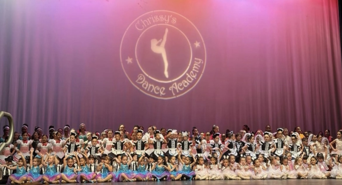 Large group of children on stage in colorful costumes, with a dance academy logo projected on purple curtain background.