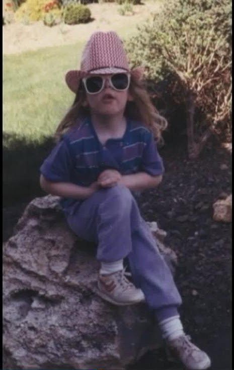 Child sitting on a rock outdoors, wearing sunglasses, a pink hat, and casual clothing, with greenery in the background.