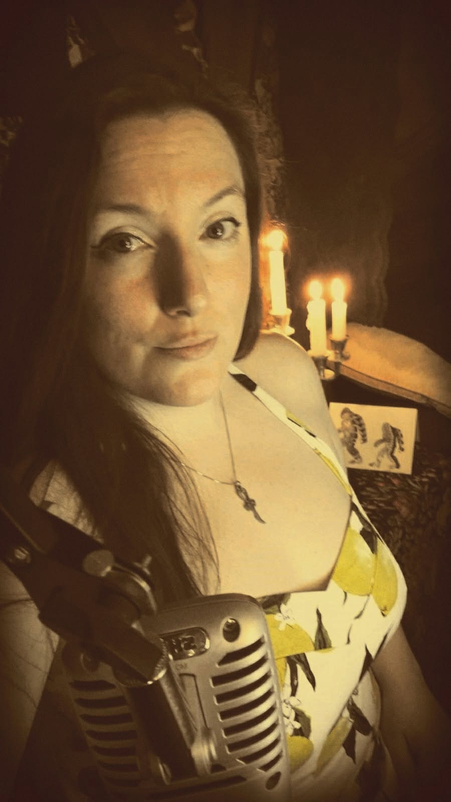 Woman with long dark hair in a patterned dress, standing near candles and a framed picture, in a dimly lit room.