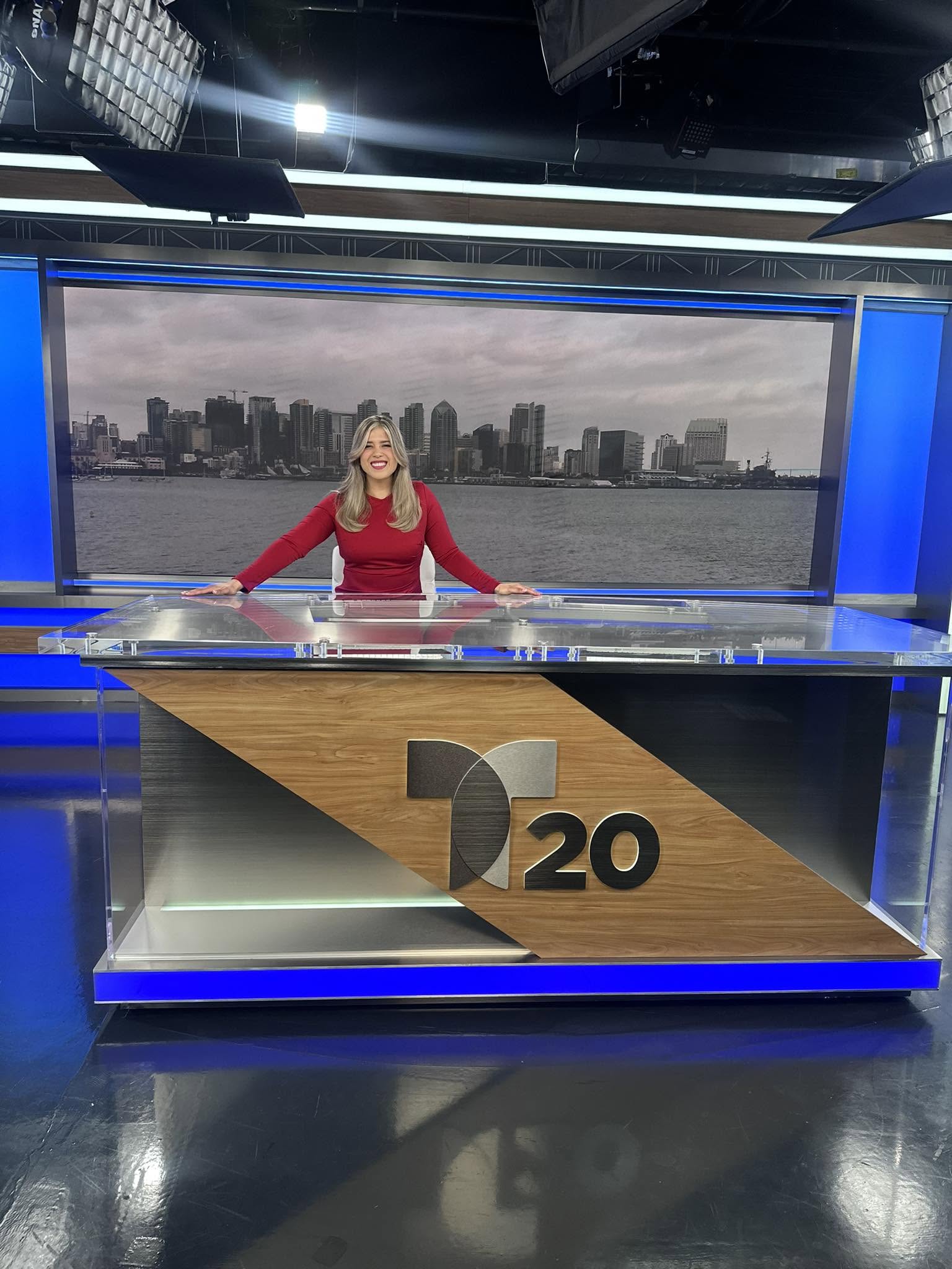 News anchor woman standing behind a desk with a cityscape background on a large screen.