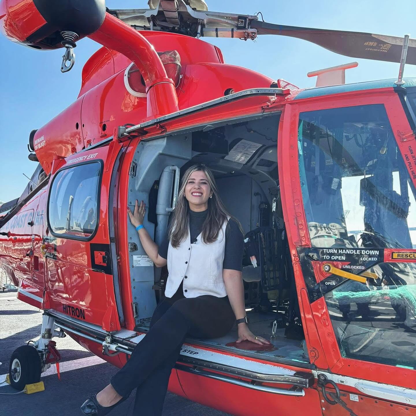 Woman sitting inside a red helicopter, smiling, with the door open and the helicopter's rotor visible above.