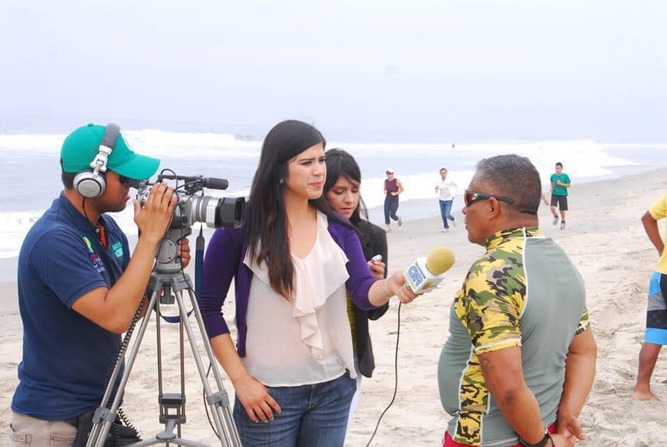 A woman interviews a man on a beach with a camera crew, others in the background, overcast sky.