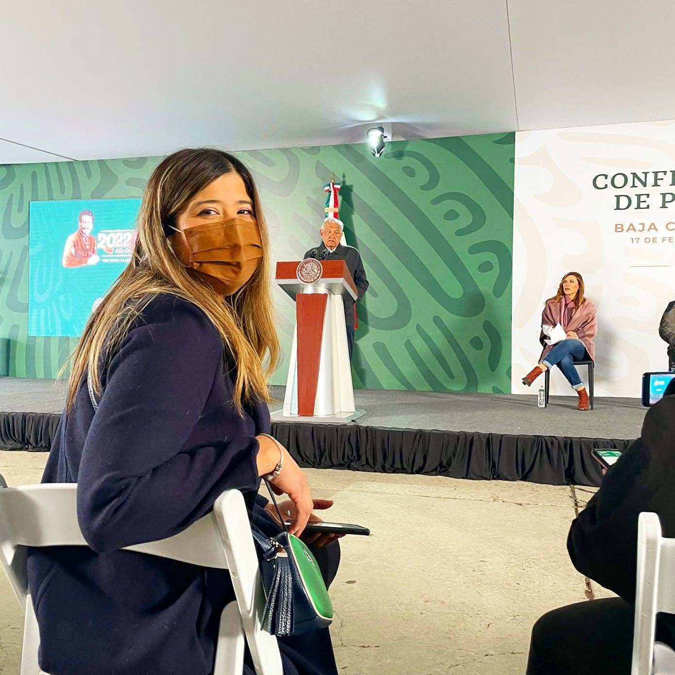 Woman with long hair wearing a brown face mask and dark clothing, sitting in a conference room with a stage and a seated woman in the background.