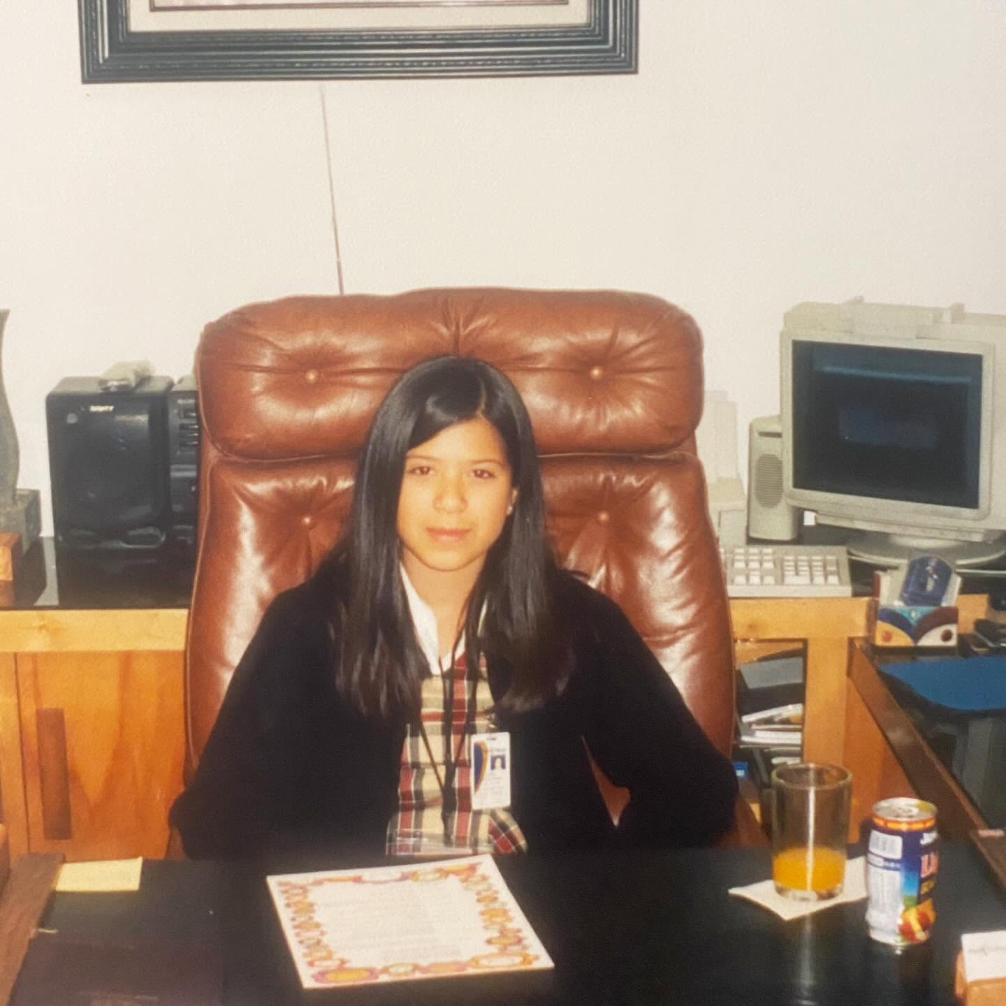 Girl with long dark hair sitting in a brown leather chair at a desk with a computer and drinks.
