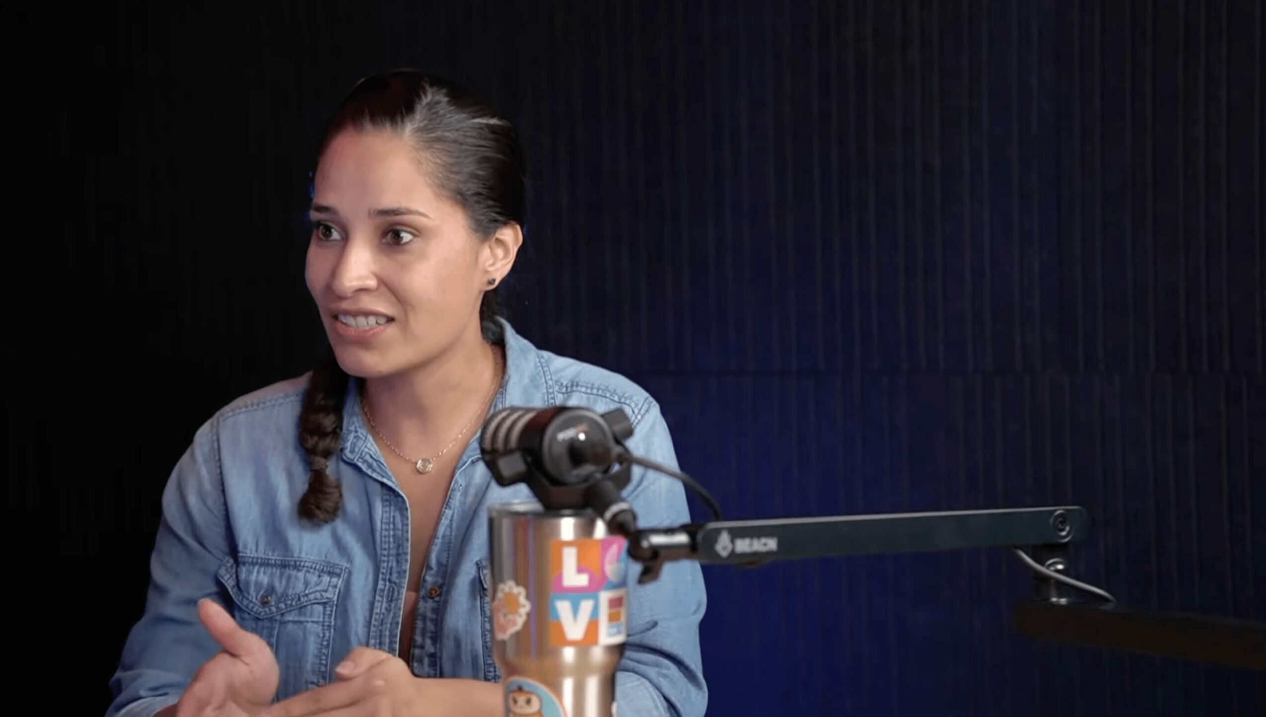 Woman with braided hair in a denim shirt speaking into a microphone, with a colorful water bottle nearby.