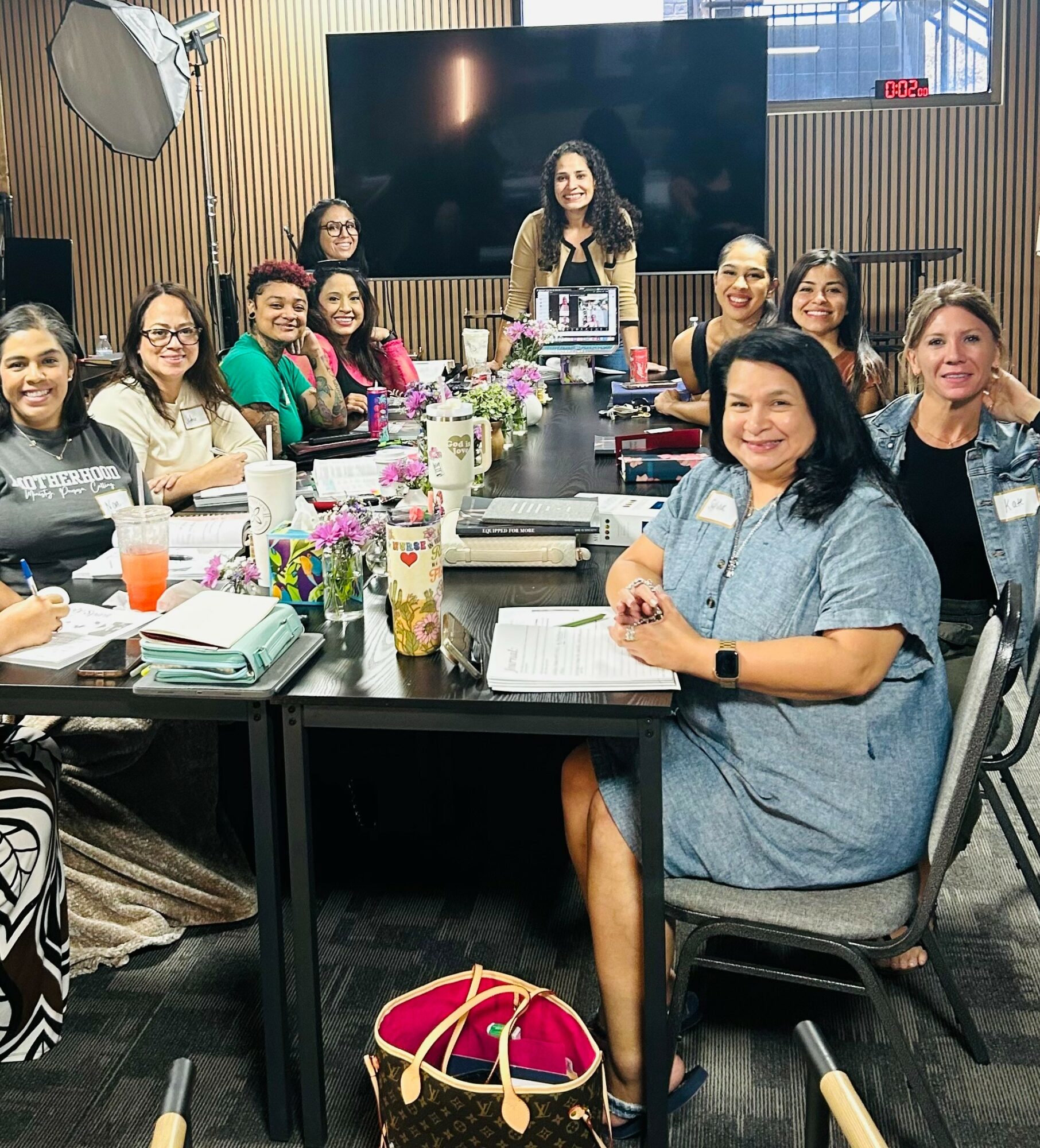 Group of women sitting around a conference table with flowers and notebooks, smiling, in a modern room with wood paneling and a large screen.