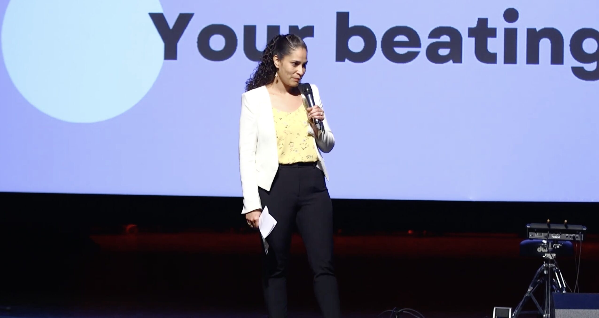 Woman speaking on stage with a large screen behind her displaying text, holding a microphone, wearing a white blazer and dark pants.