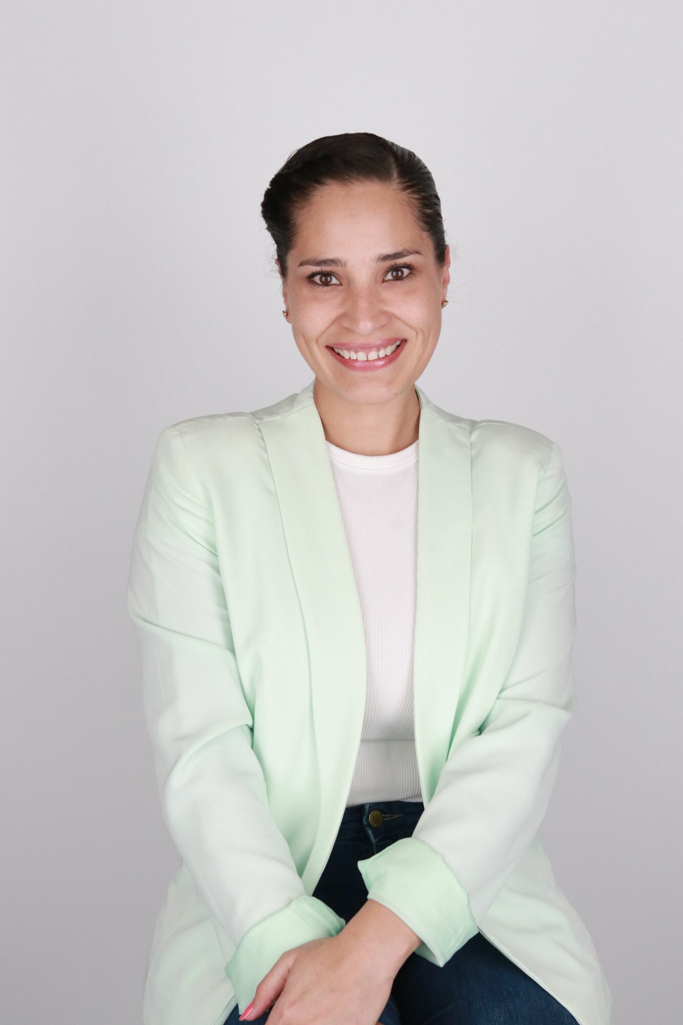 Woman with dark hair in a ponytail smiling, wearing a light green blazer and white top, standing against a plain background.