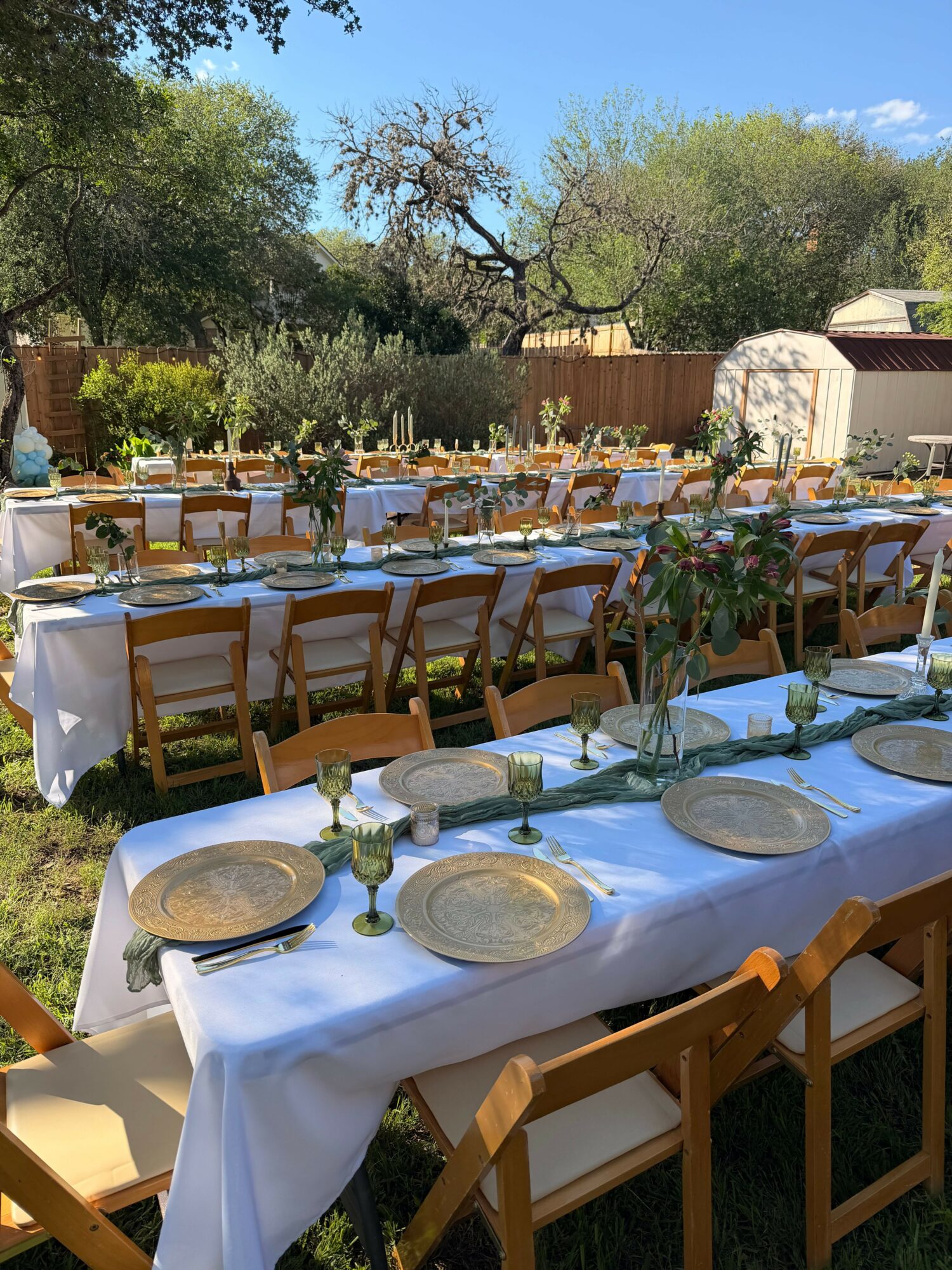 Long outdoor table set with plates, glasses, and chairs in a garden with trees and a fence, sunny sky.