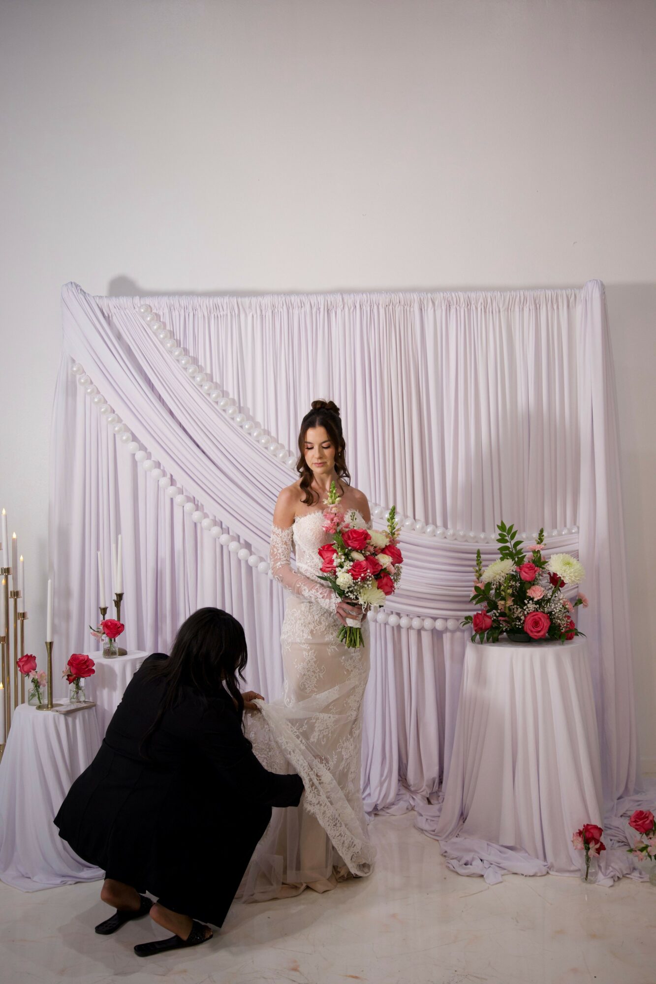 Woman in wedding dress holding bouquet, woman kneeling adjusting dress, decorated backdrop with flowers and drapes