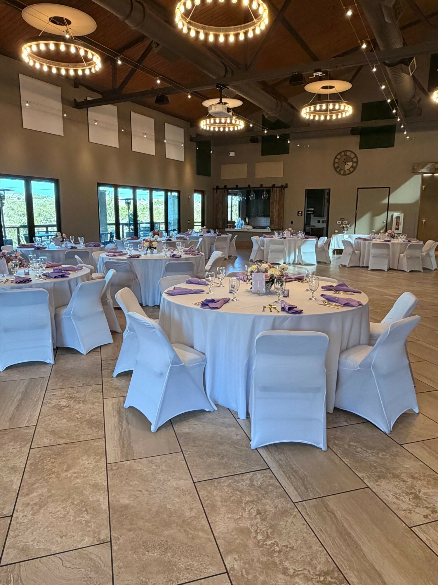 Event hall with round tables covered in white cloths, chairs with white covers, and large windows, decorated for a celebration.