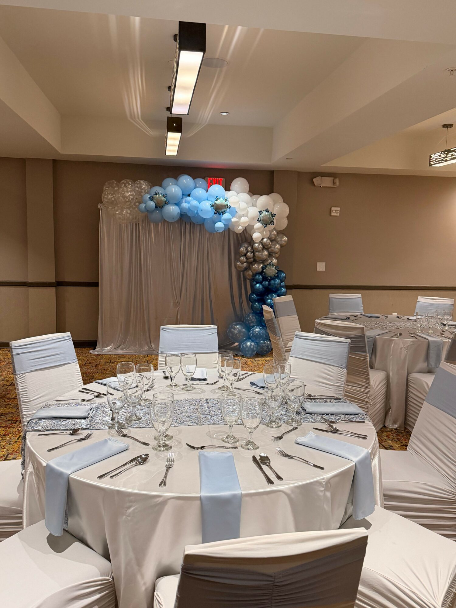 Round table with white tablecloth, set with glasses and silverware, decorated for an event with balloon arrangement in blue, white, and silver, and a beige backdrop.