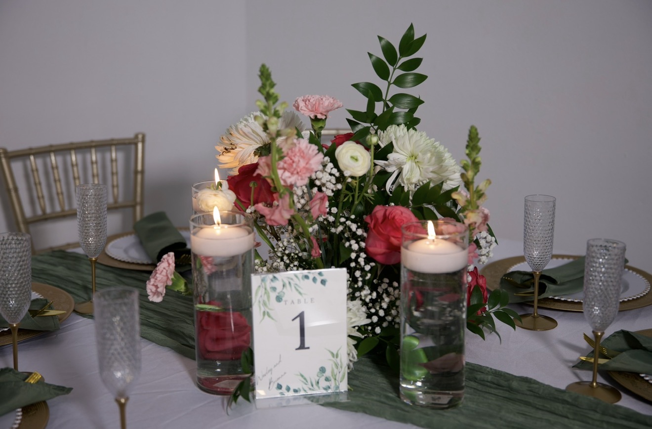 Table centerpiece with pink and white flowers, candles, and a table number card, set for a formal event.