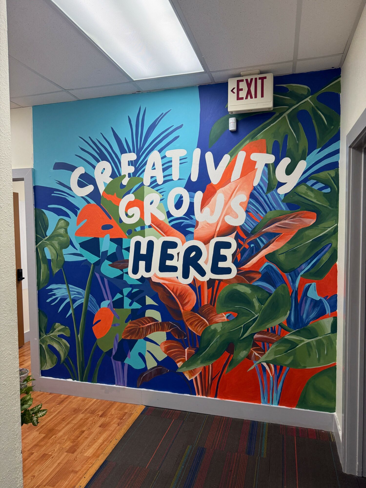 Colorful wall mural with tropical plants and the text 'Creativity Grows Here' in bold letters, near an exit sign.