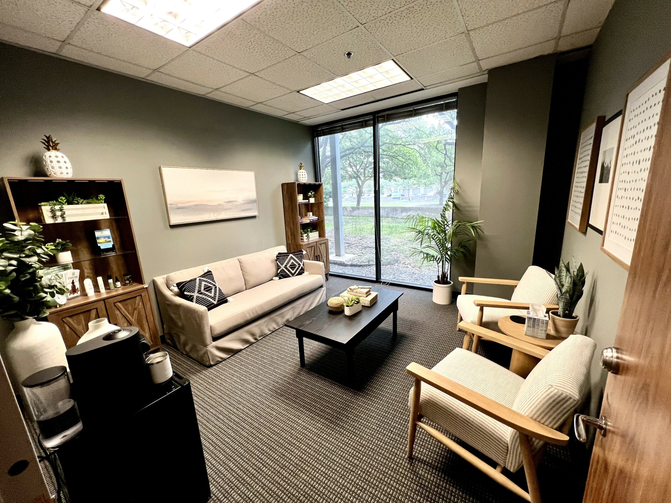 Office waiting area with sofa, two armchairs, coffee table, bookshelf, and large window with greenery outside.