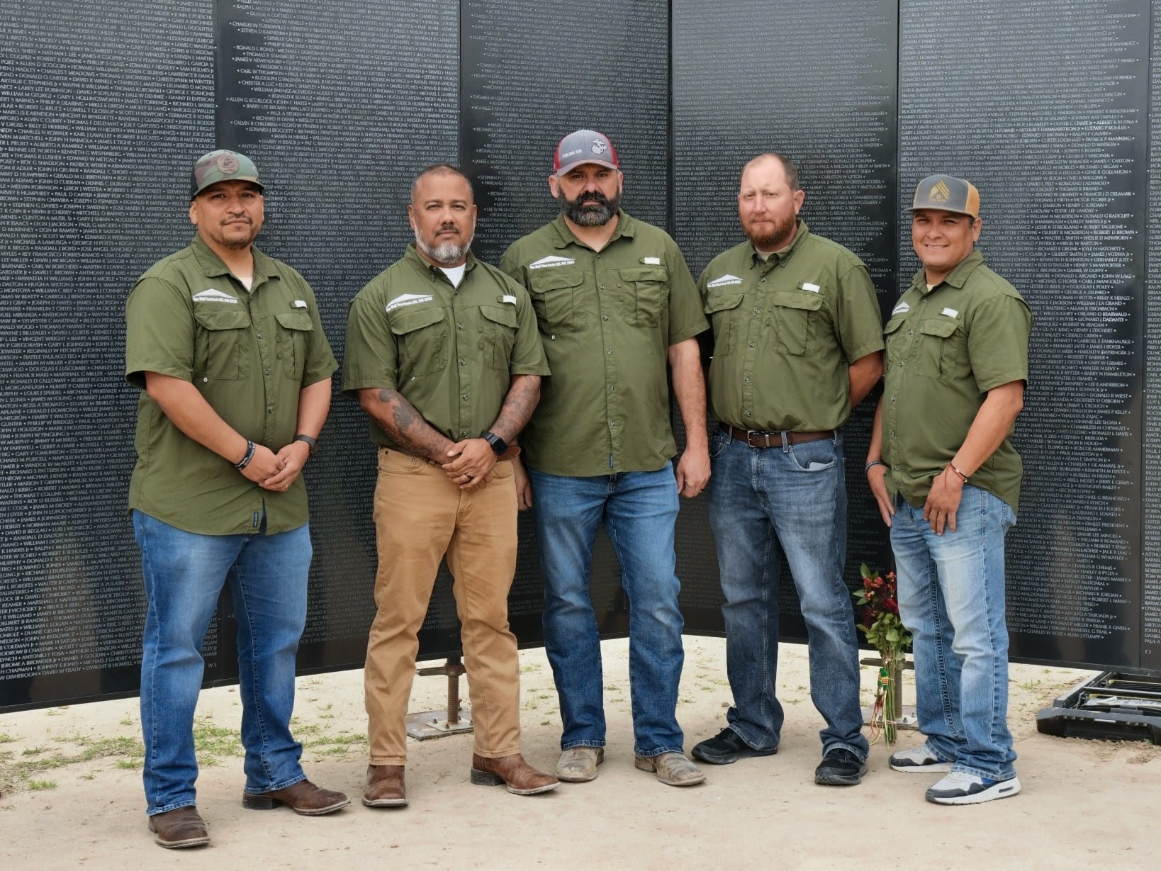 Five men stand outdoors in front of a dark wall, wearing green shirts and casual pants, posing for a group photo.