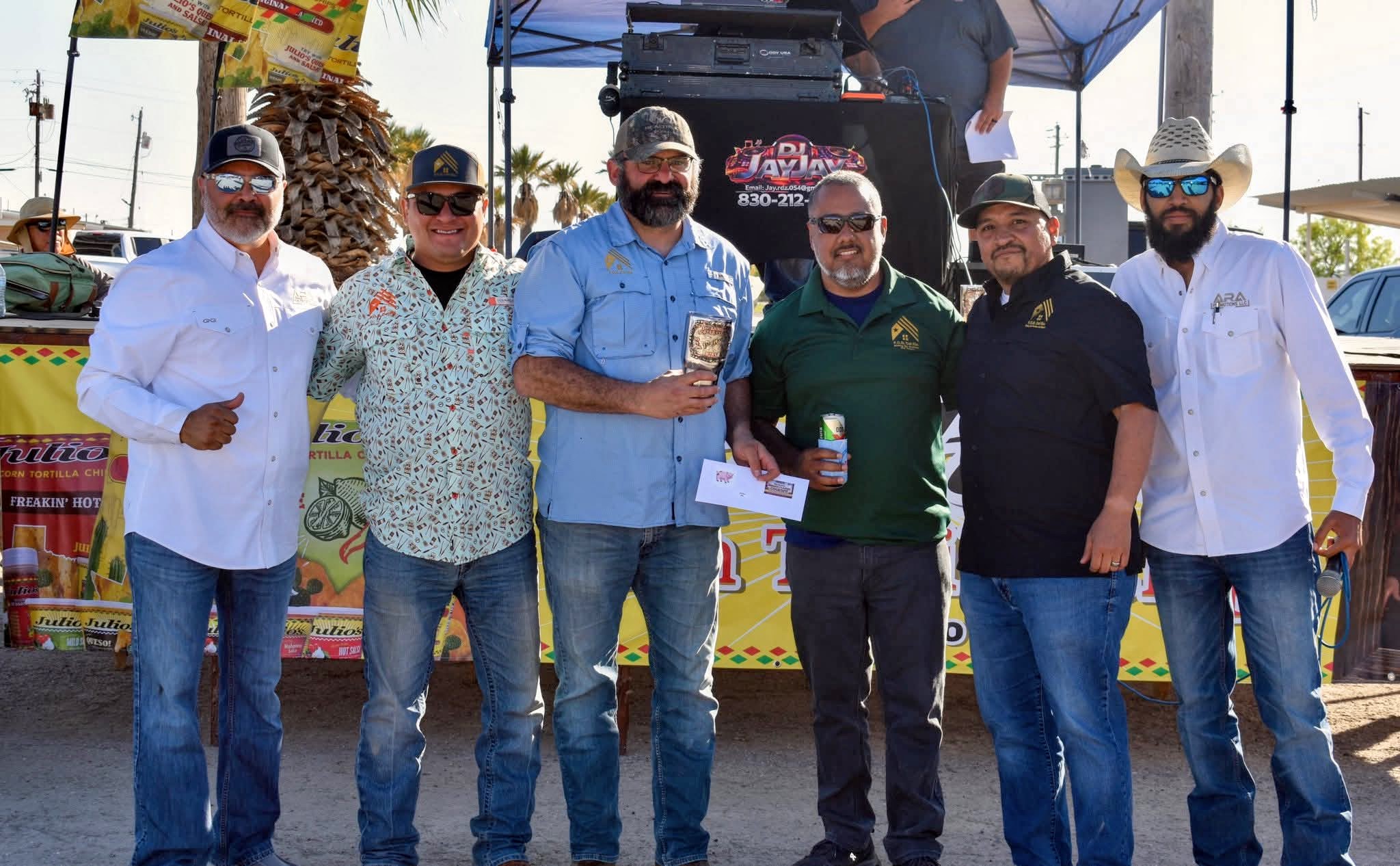 Group of six men standing outdoors, some holding drinks, in front of a colorful booth and a stage with a canopy.