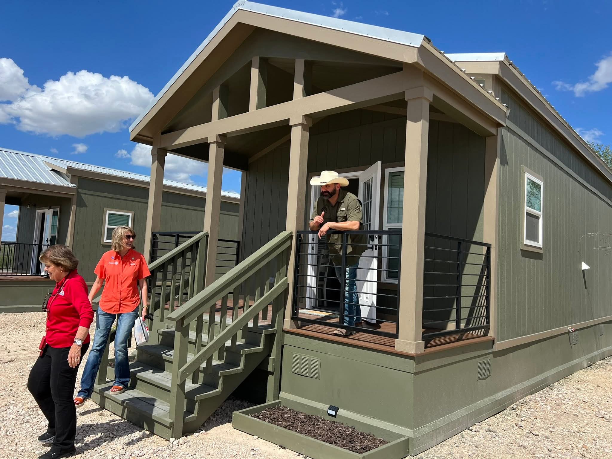 People standing on front porch of a house with stairs, railing, and a person wearing a hat near the door.