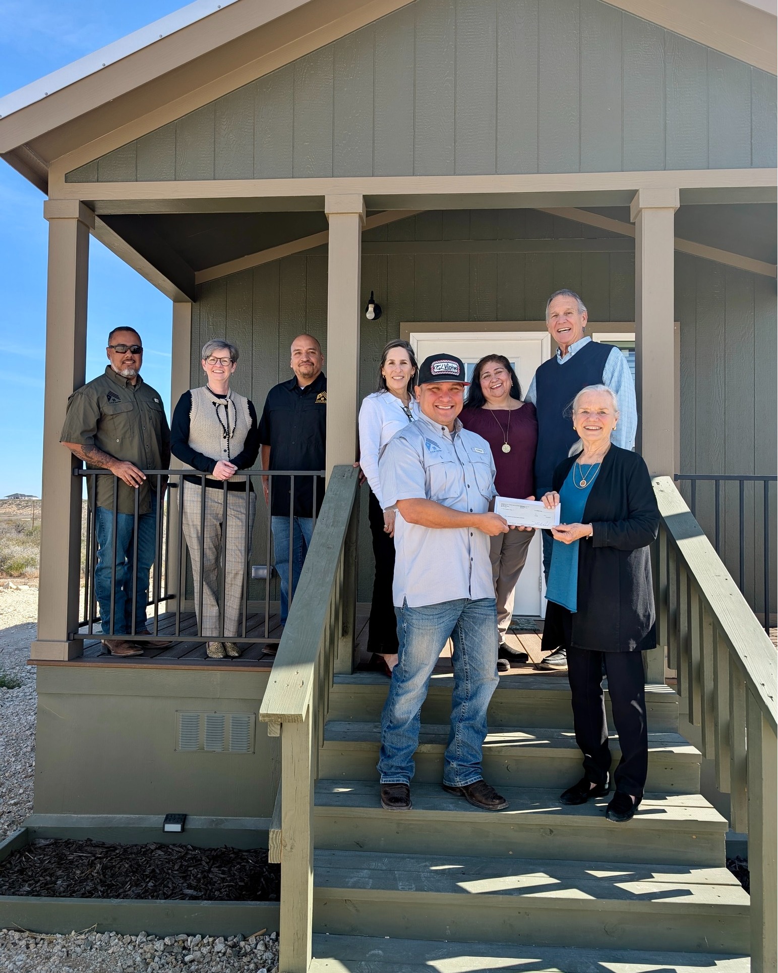 Group of seven people standing on porch steps in front of a house, some holding a document, smiling.