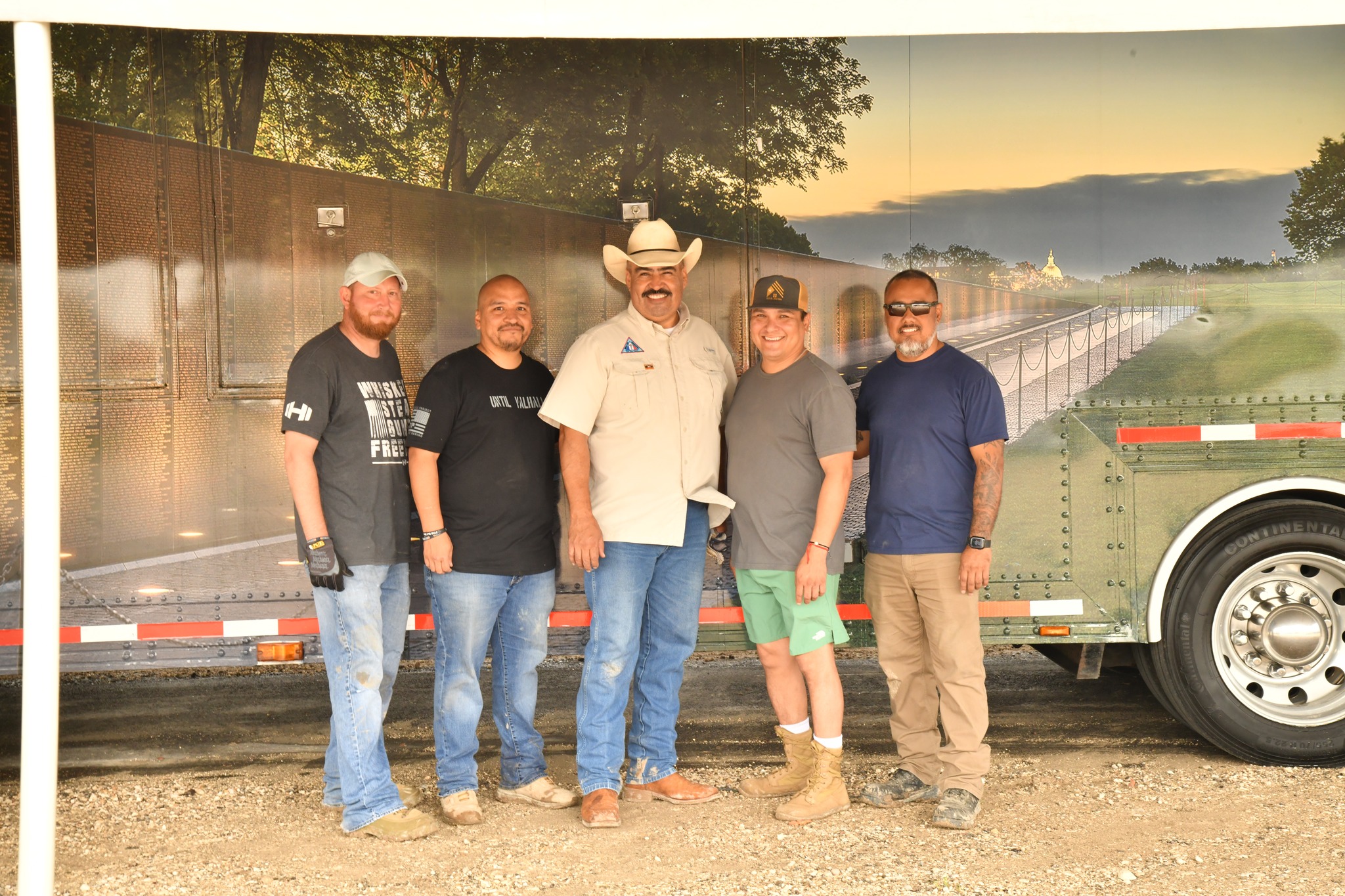 Five men standing outdoors in front of a large vehicle, with a fence and trees in the background, during sunset.