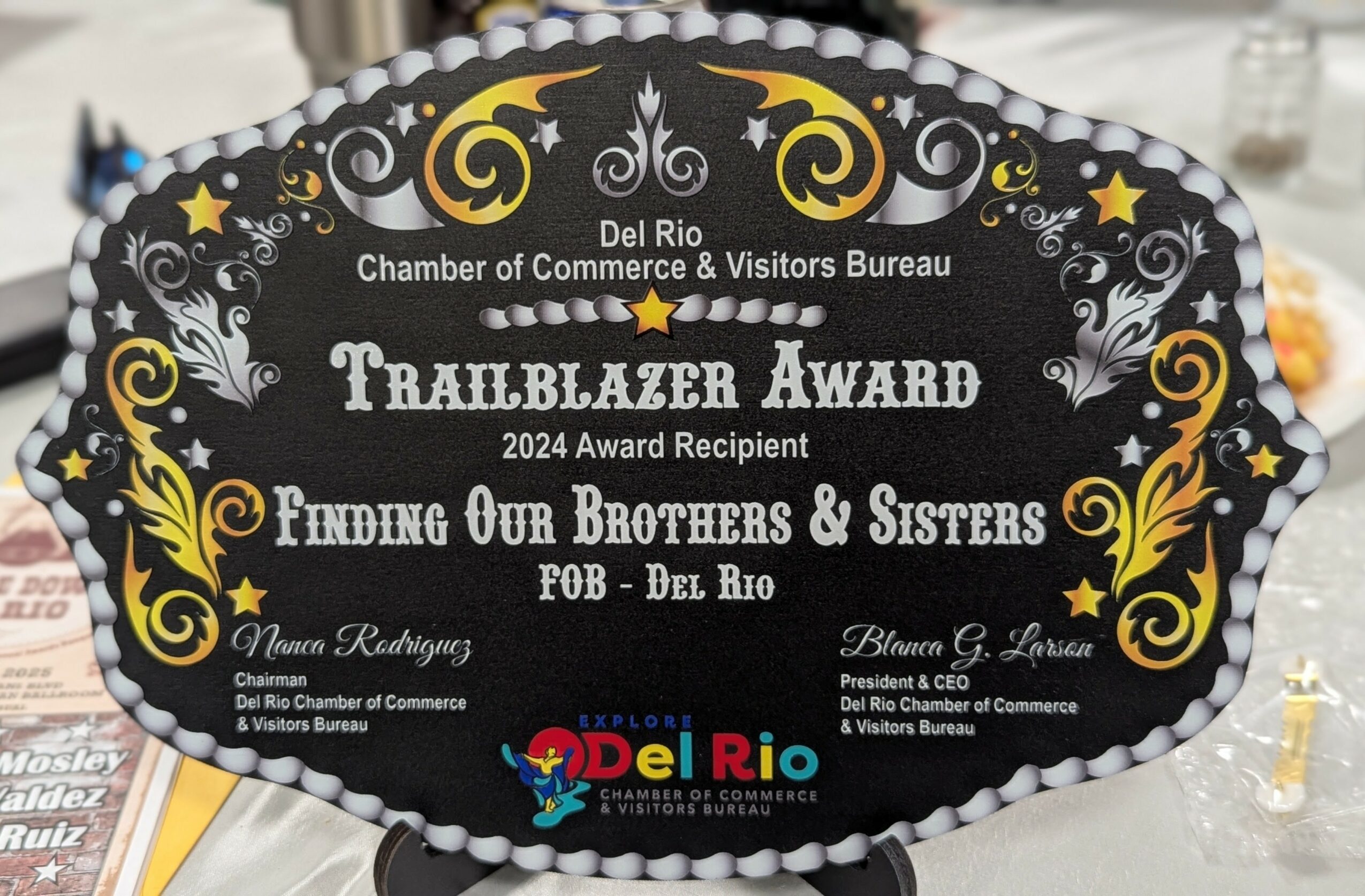 Black decorative award plaque with gold and white accents, text about the Trailblazer Award, and logos at the bottom.