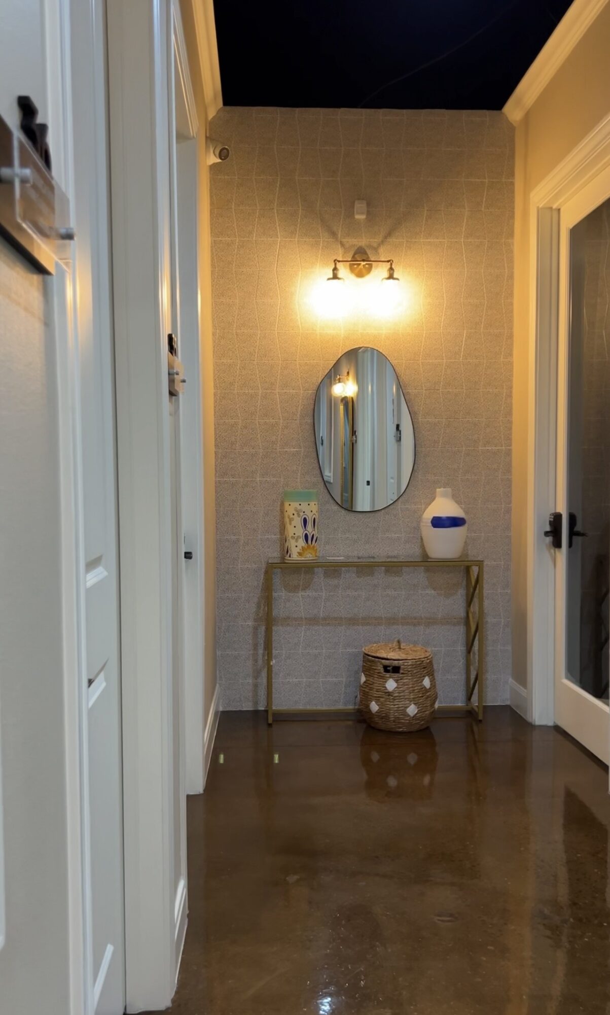 Narrow hallway with a console table, oval mirror, wall light, and decorative items, leading to a door on the right.
