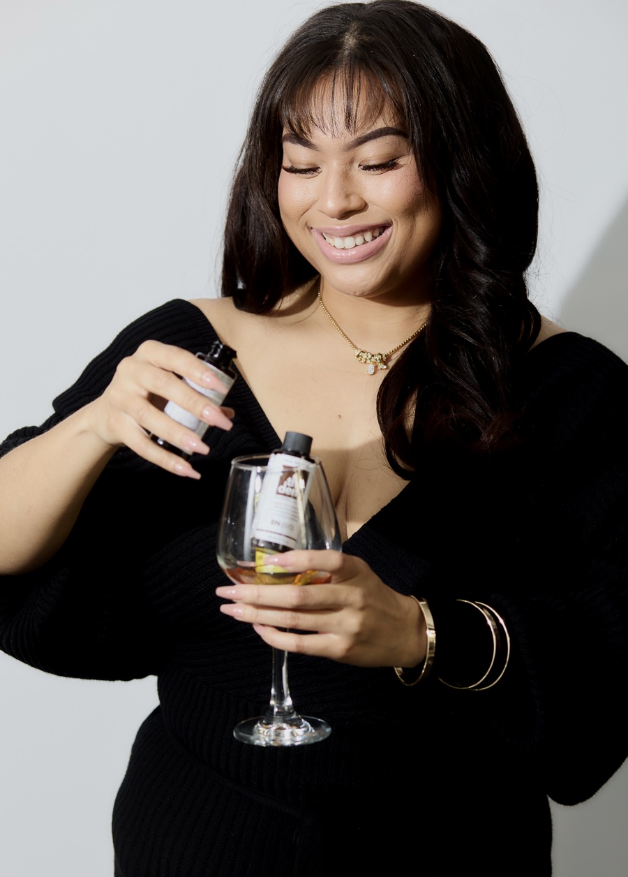 Smiling woman with dark hair holding a glass of wine, wearing a black top, against a plain background.