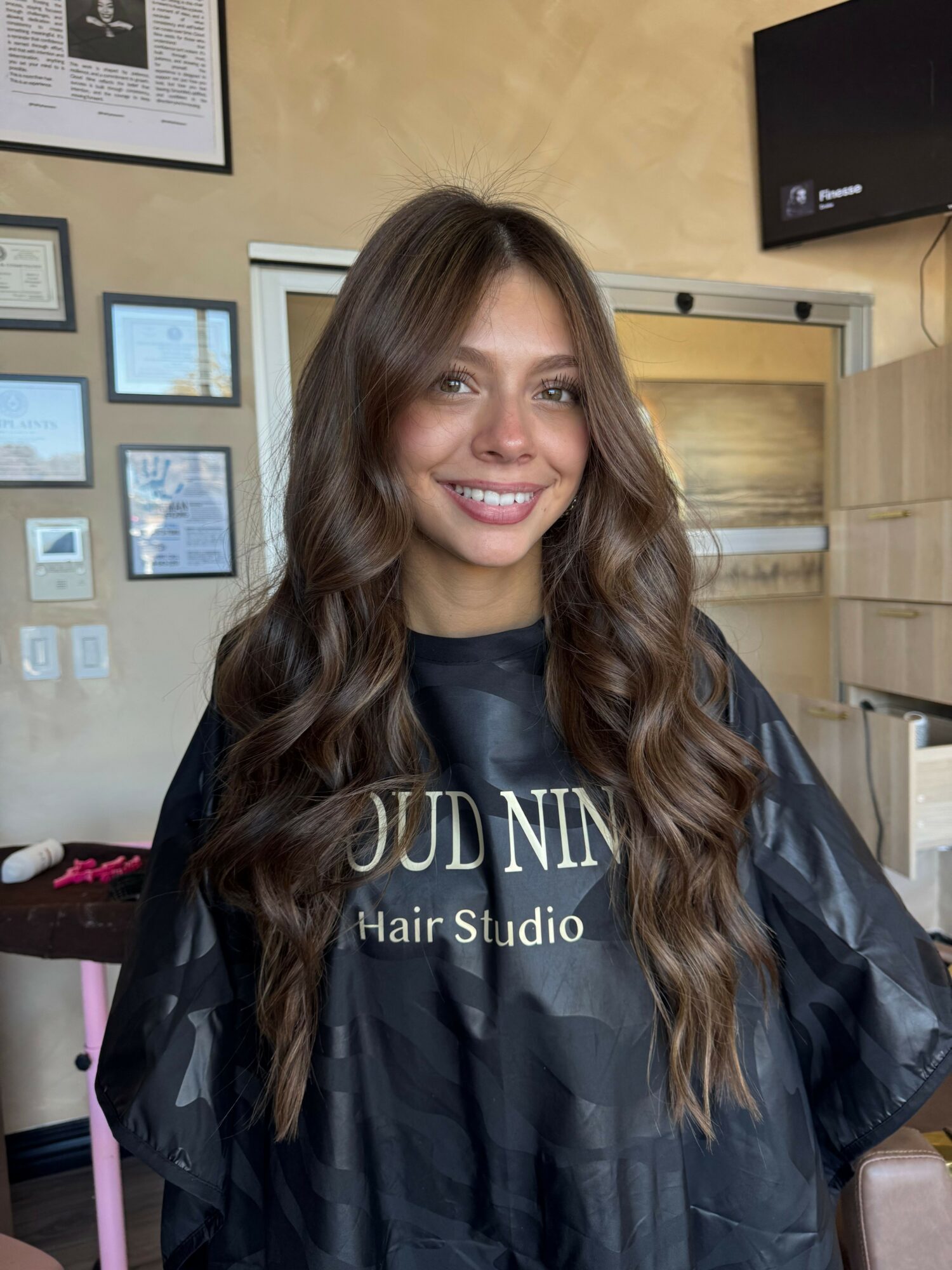 Young woman with long wavy hair smiling in a hair salon, wearing a black cape with salon logo, background with framed pictures and a TV.