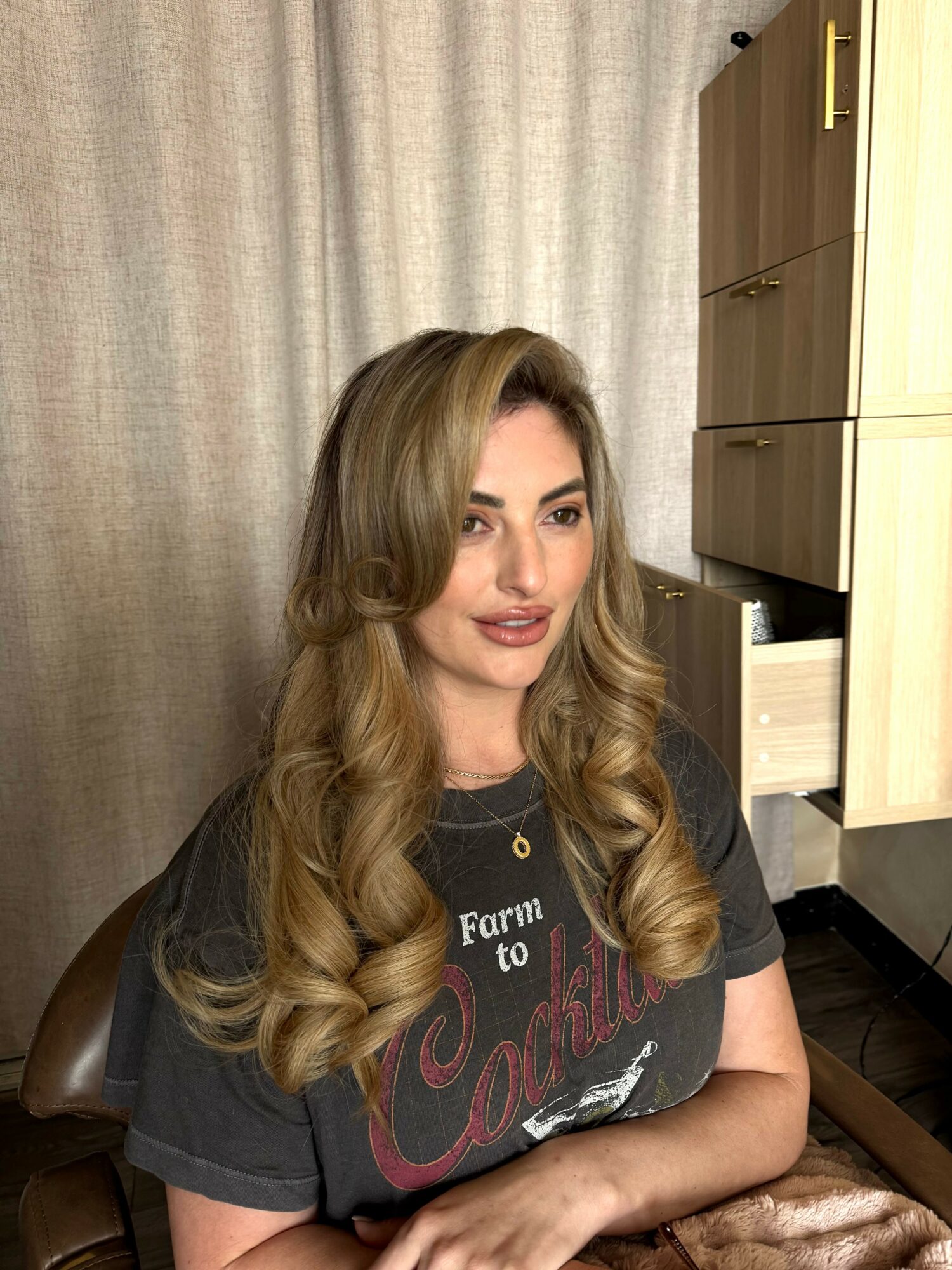Woman with long wavy hair sitting indoors, wearing a dark T-shirt, in front of a curtain and wooden furniture.
