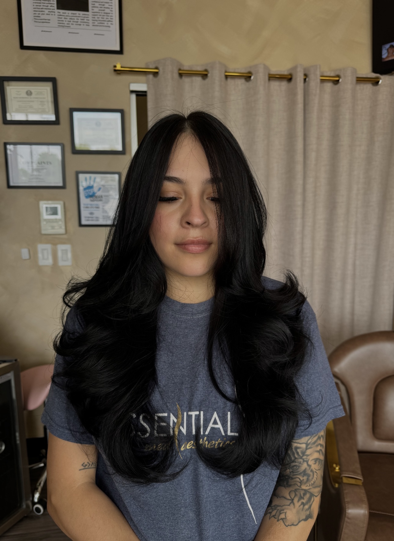 Young woman with long, wavy black hair wearing a gray T-shirt, standing indoors with framed pictures and curtains in the background.